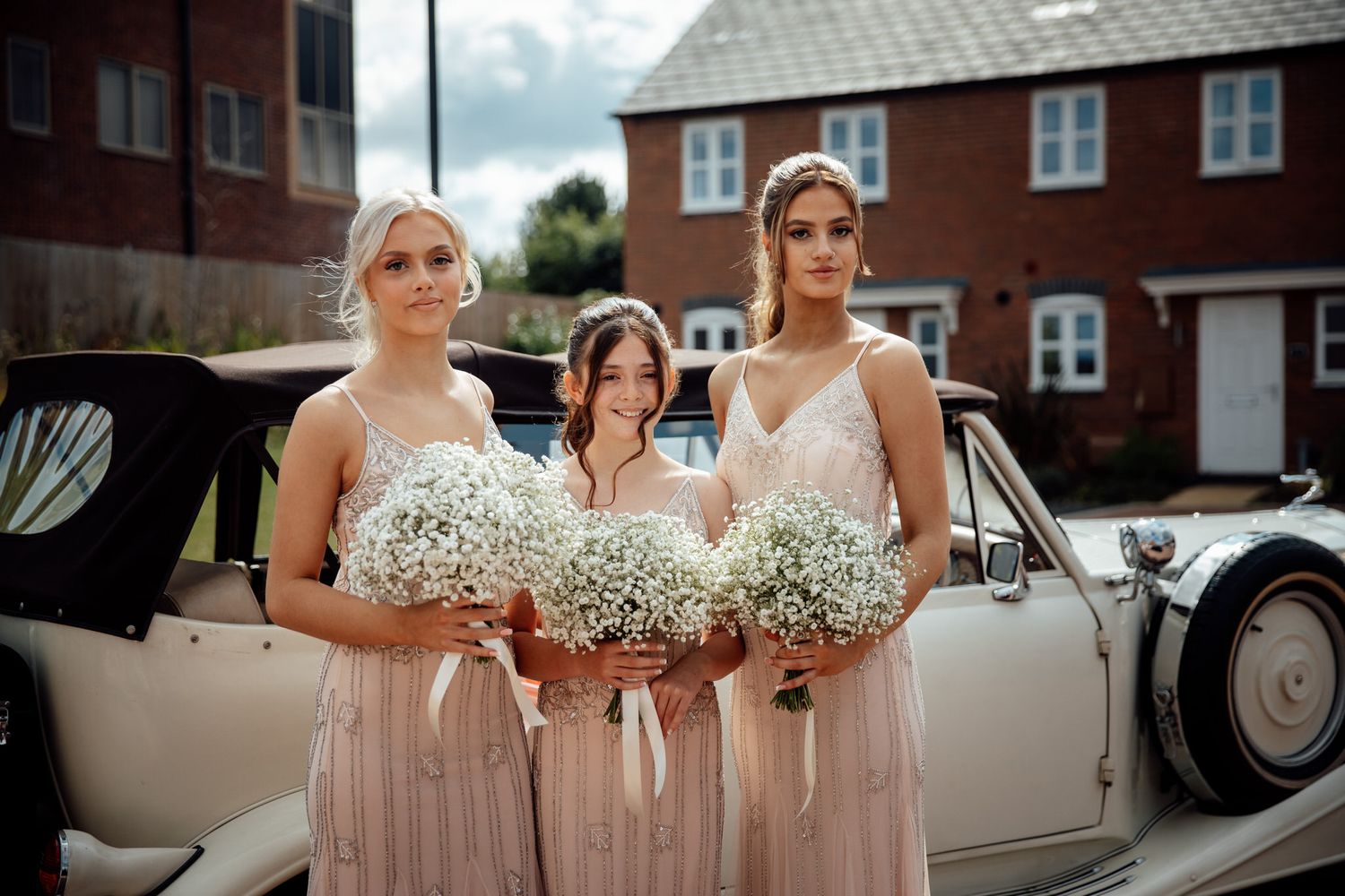 Classic vintage car parked outside brick building with wedding party in pink dresses holding white bouquets.