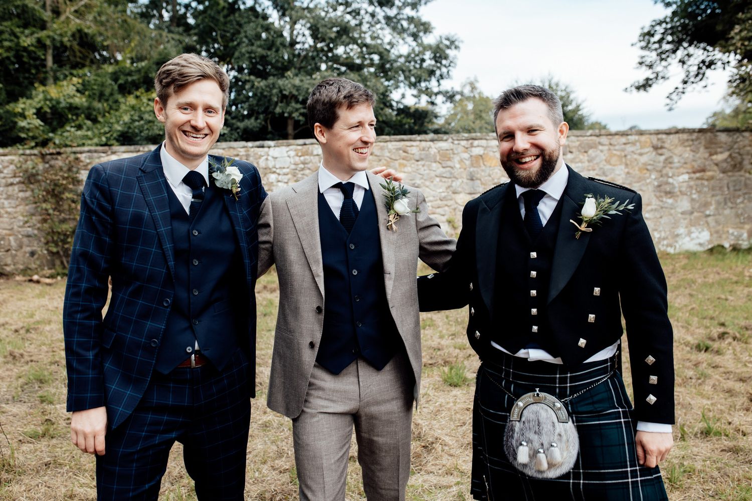 Wedding party members in formal attire gather outdoors against a stone wall backdrop.