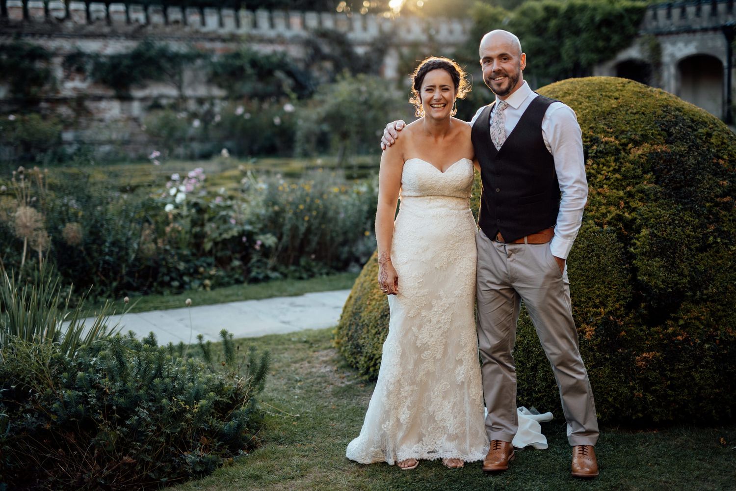 Wedding couple walks through lush garden path near historic brick building.
