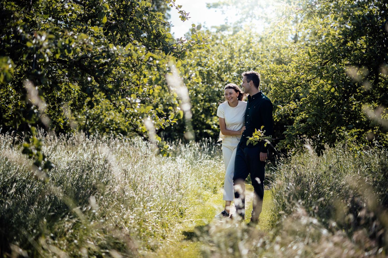 Couple walking through golden meadow with sunlit trees in background.