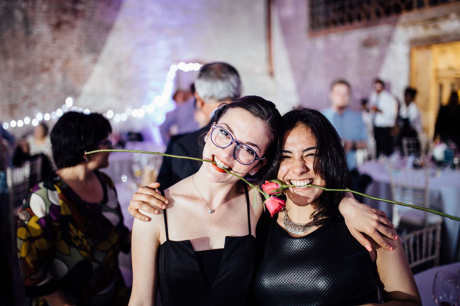 Two guests playfully pose with roses at an evening wedding reception.