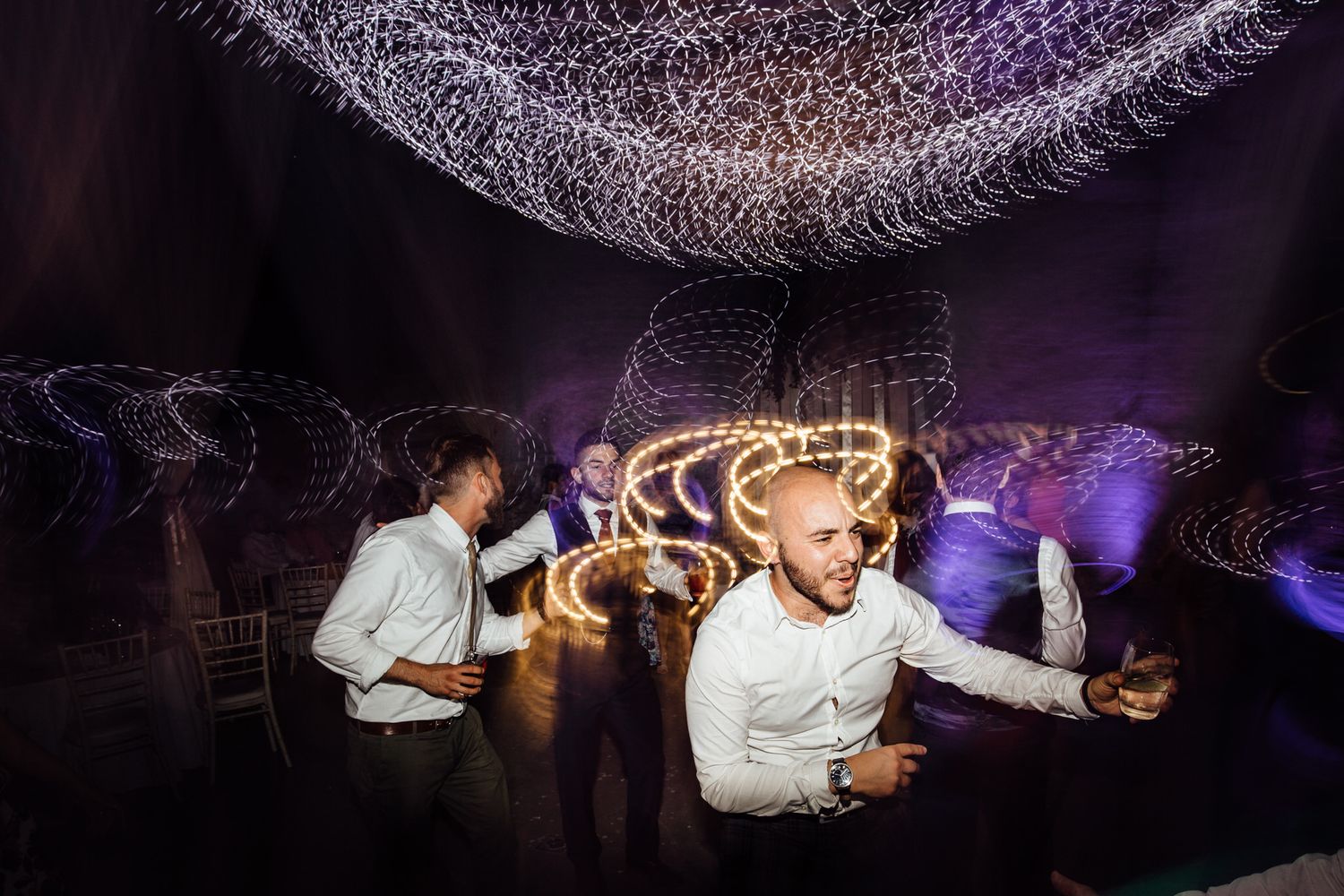 Wedding reception guests dancing in a venue with purple uplighting and white fabric decor.