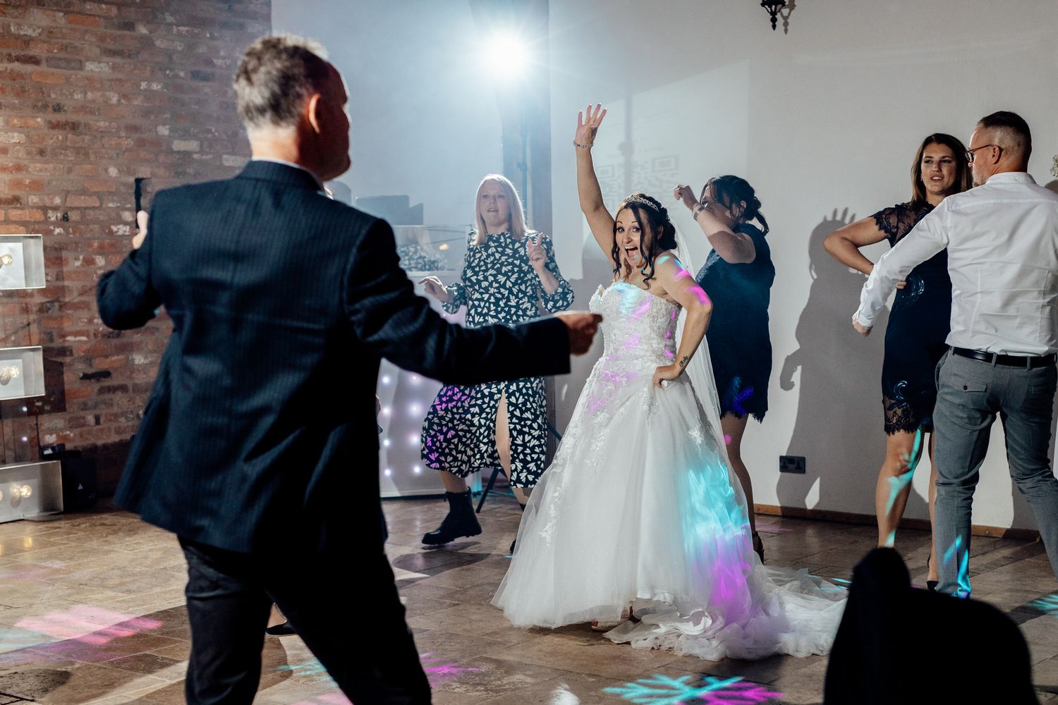 Wedding reception dance party with guests celebrating under disco lights in brick-walled venue.