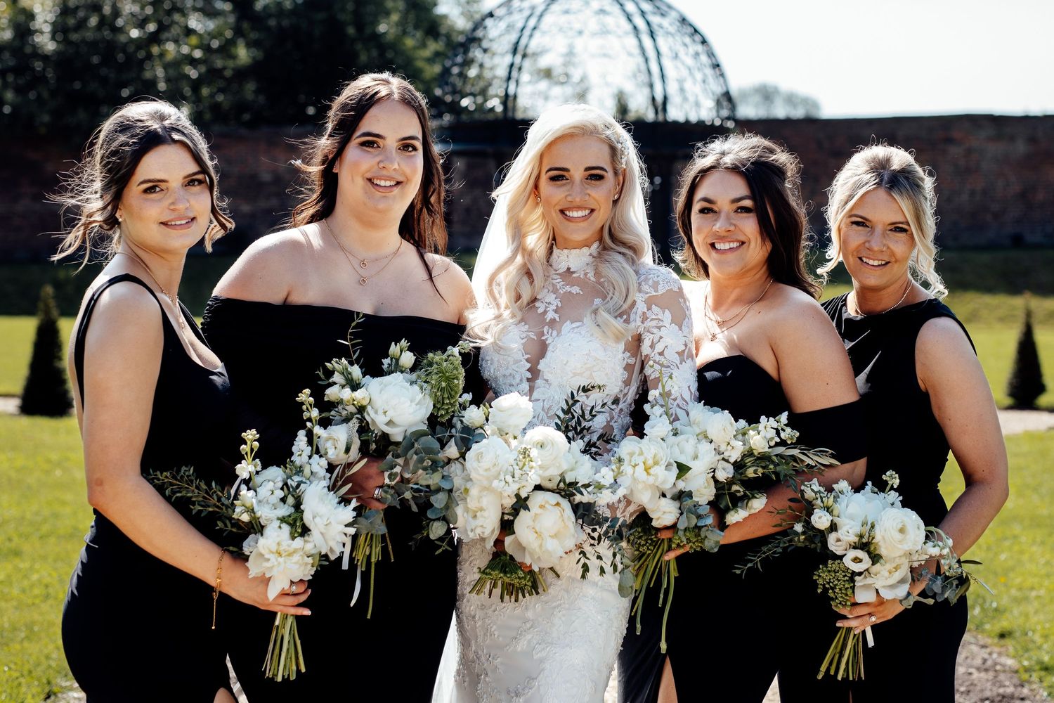 Bridesmaids in black dresses holding white floral bouquets pose together outdoors on a sunny day.