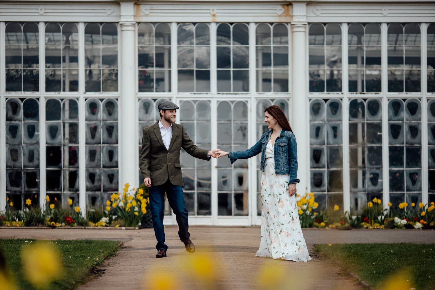 Elegant couple poses in front of a grand conservatory with blooming flowers.