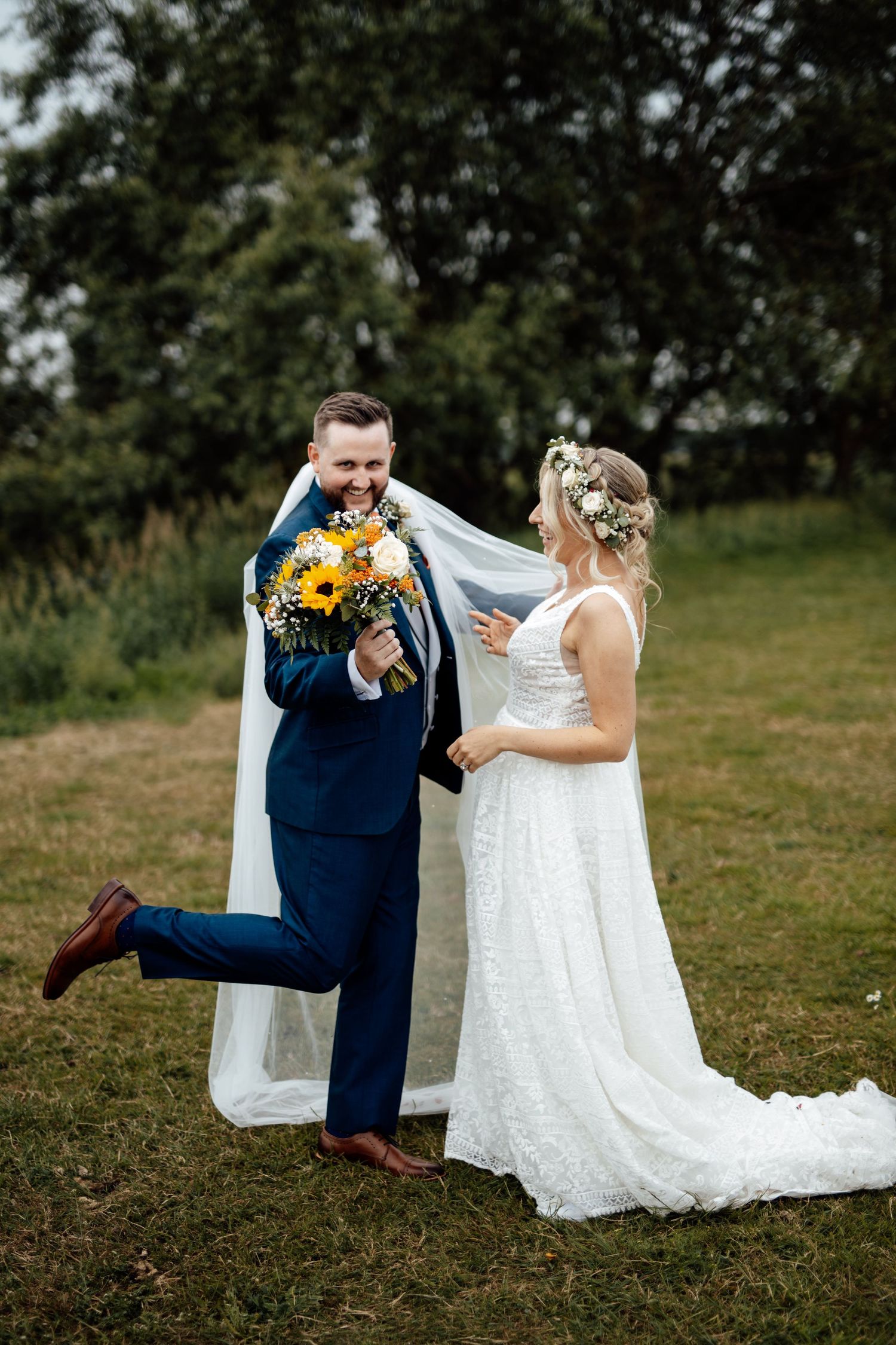 Couple in wedding attire sharing a joyful moment on grass with navy suit and white dress details.