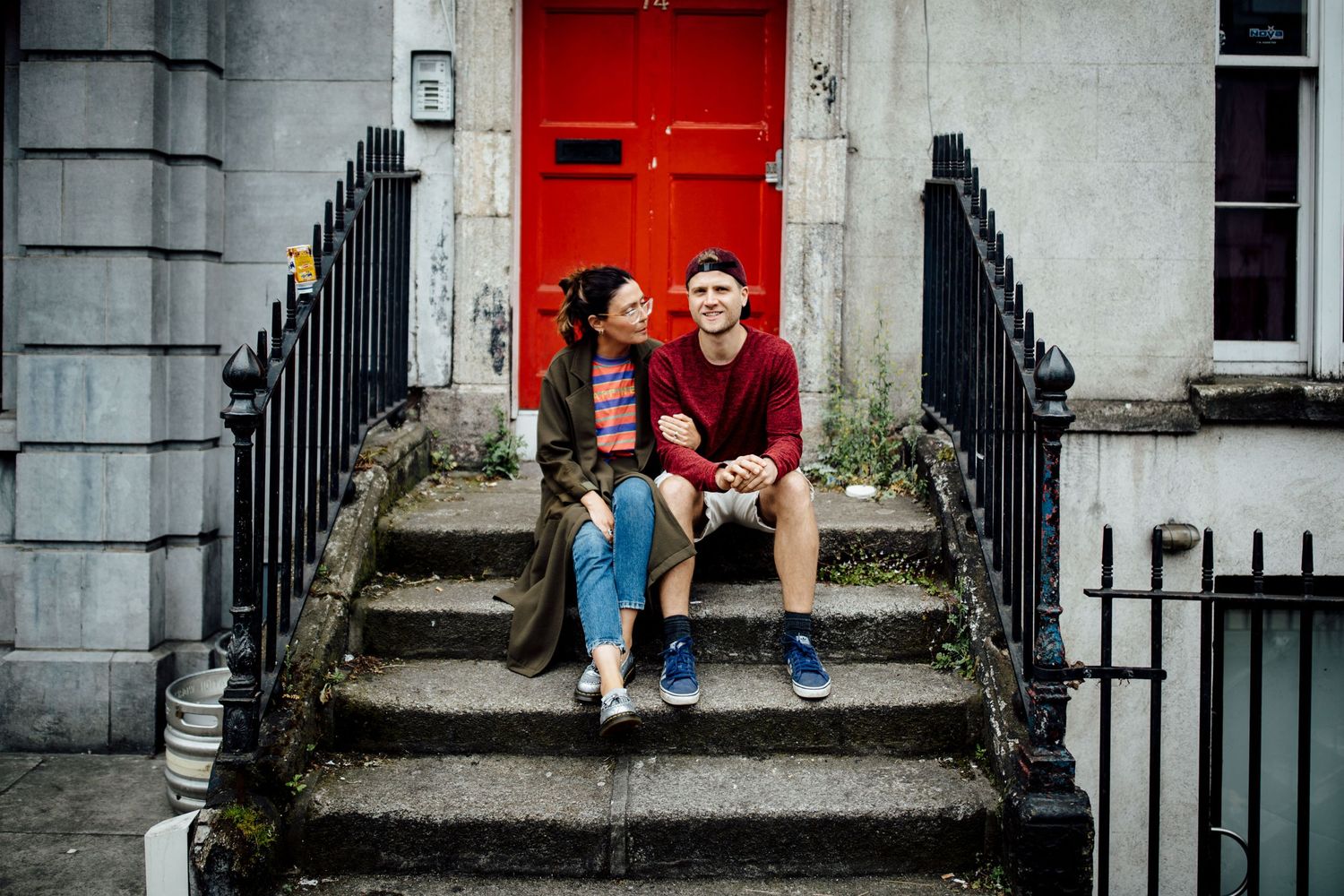 A couple sits on stone steps in front of a classic red door and iron railings.