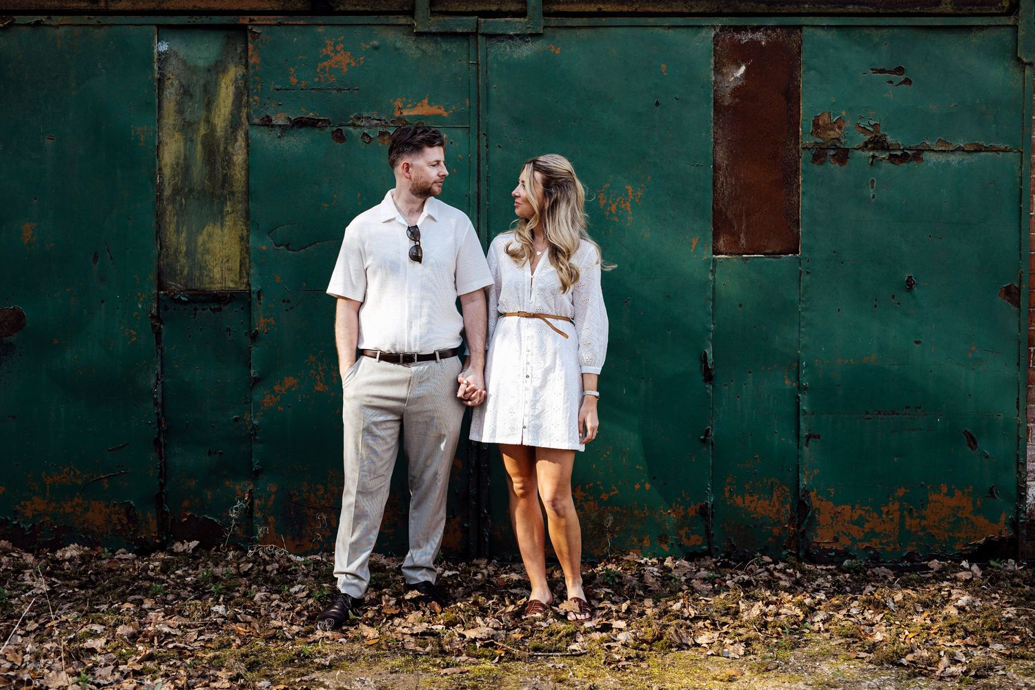 Romantic couple in white outfits pose against rustic teal wall during outdoor photoshoot.