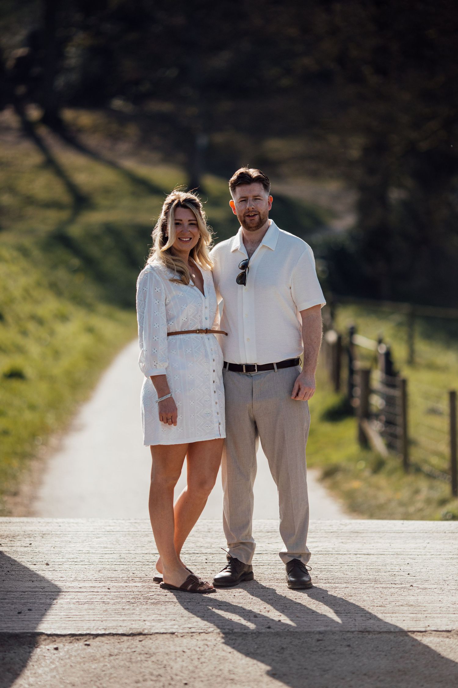 Two people in coordinated white outfits walking along a sunlit path with wooden fencing.