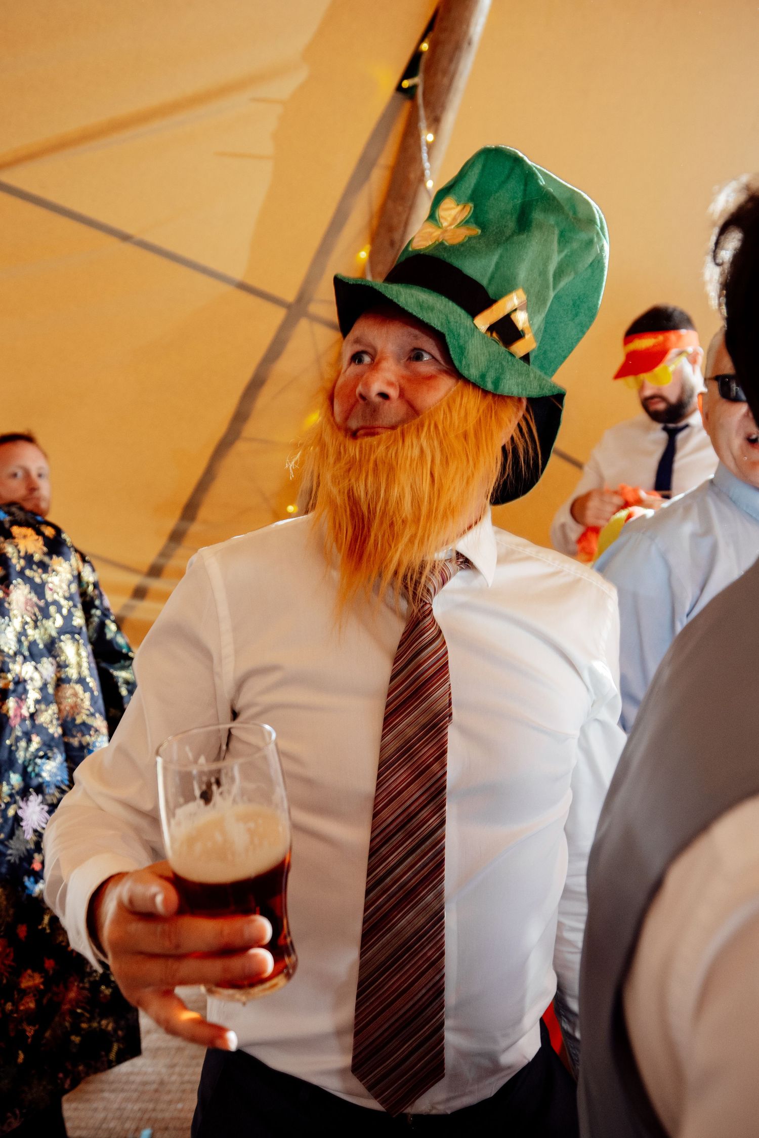 Wedding guest in leprechaun costume with fake orange beard holds a beer at reception.