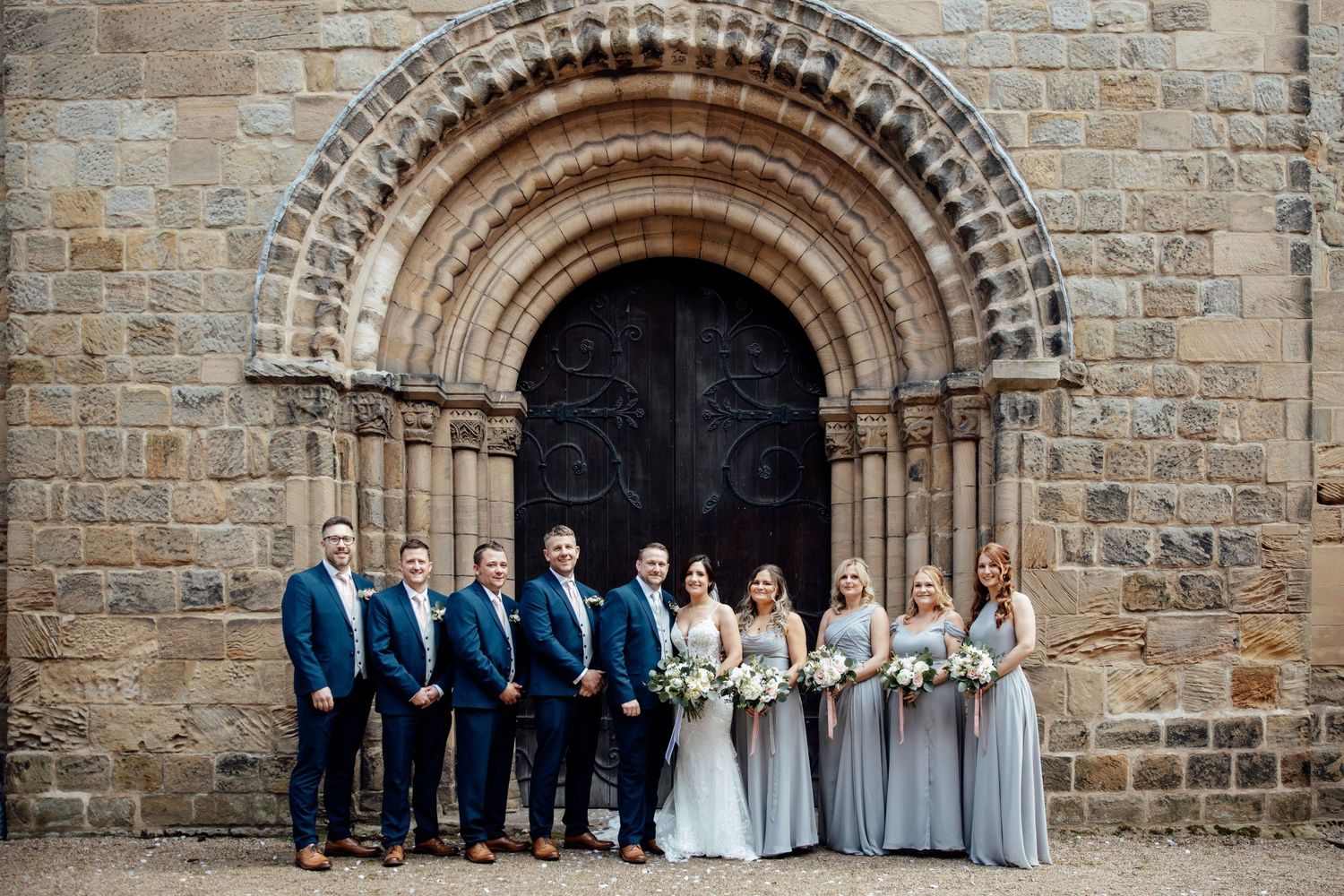 Wedding party poses in front of stone church archway for formal group photo.