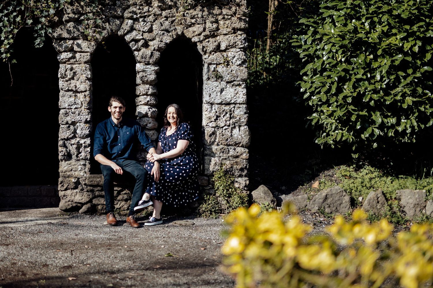 A couple sits in a stone archway with yellow flowers blooming in the foreground.