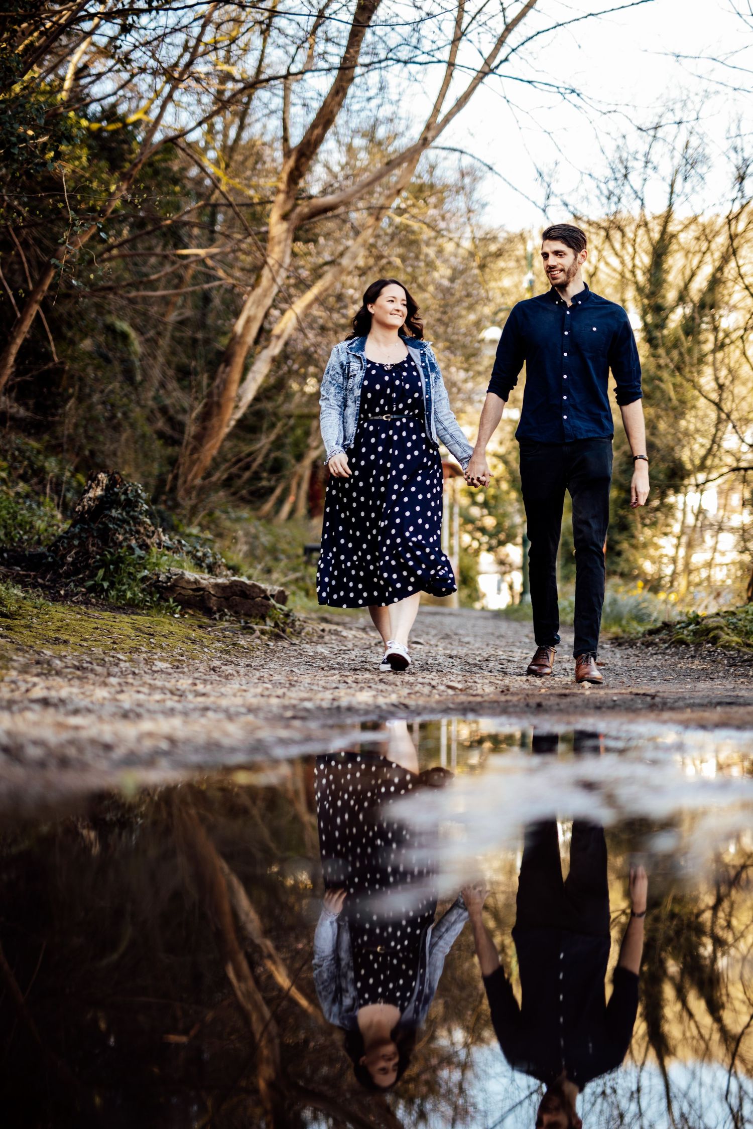 A couple's reflection appears in a puddle as they walk together on an autumn day.