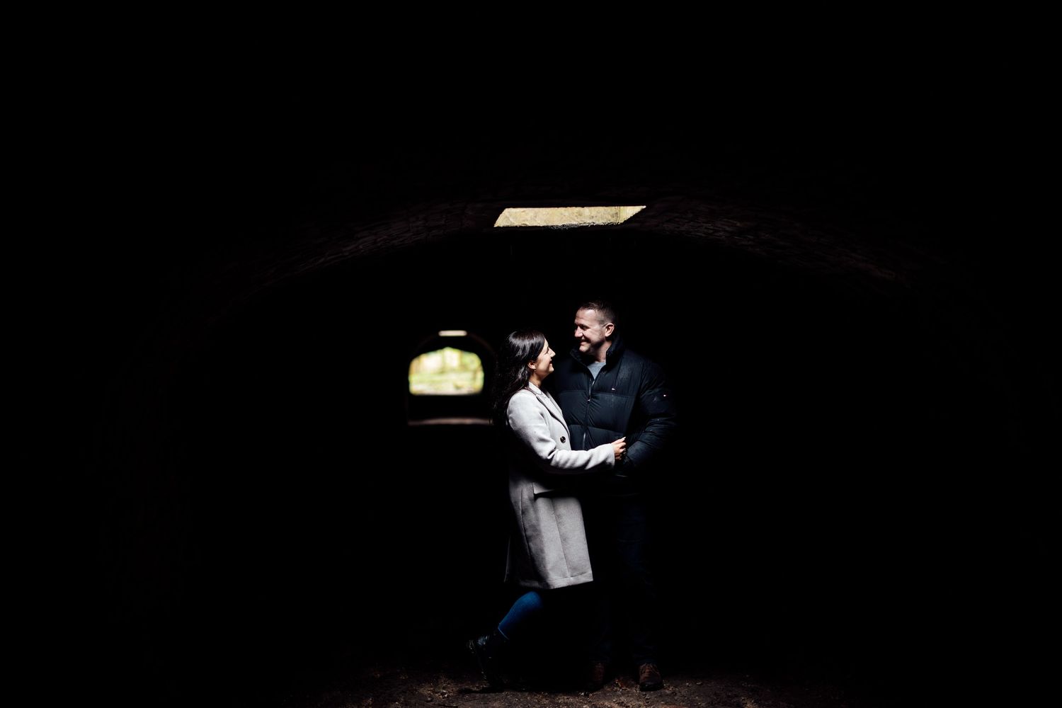 A couple walks through a dimly lit hallway holding hands and umbrellas.