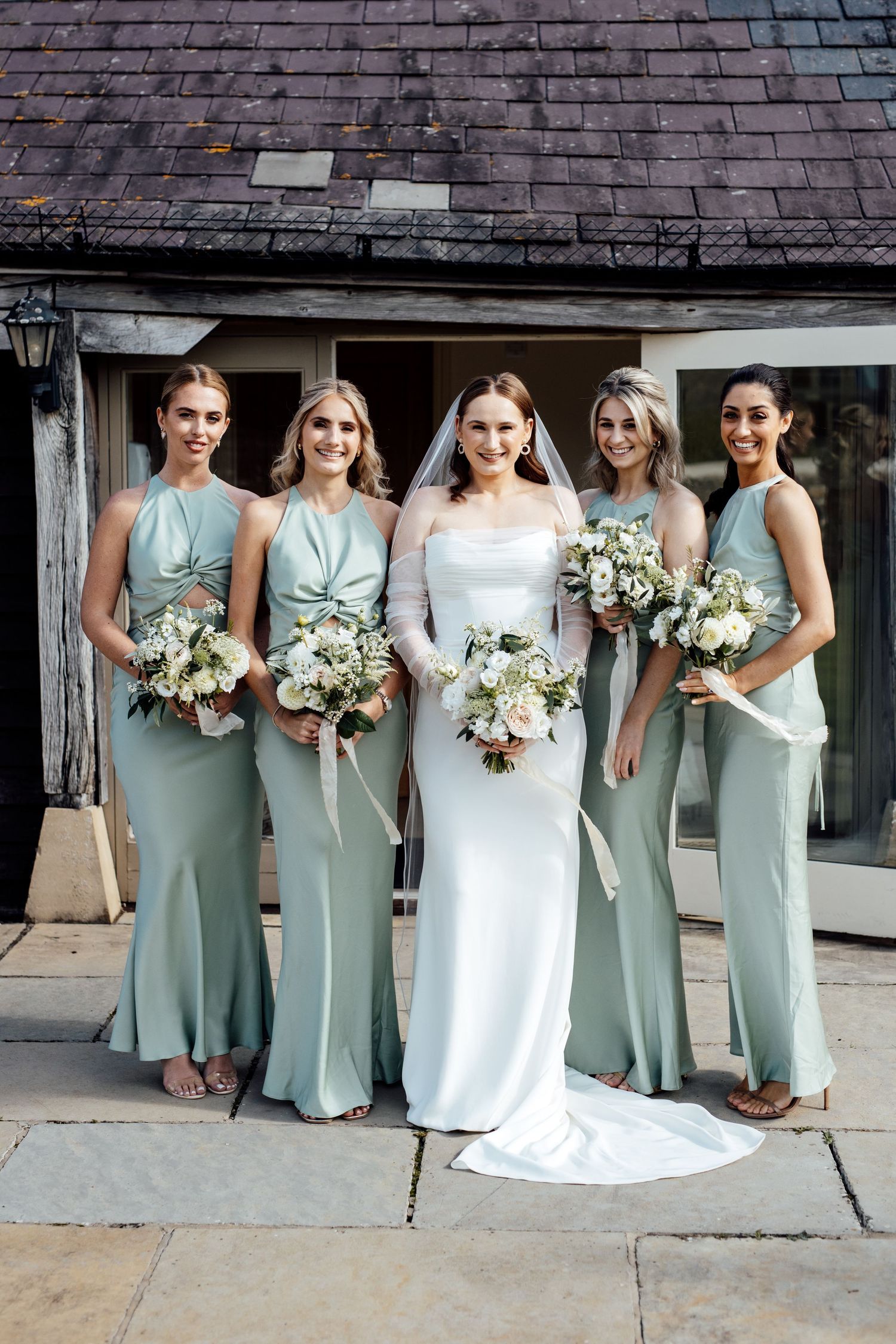Bridesmaids in sage green dresses pose with the bride outside a historic building.