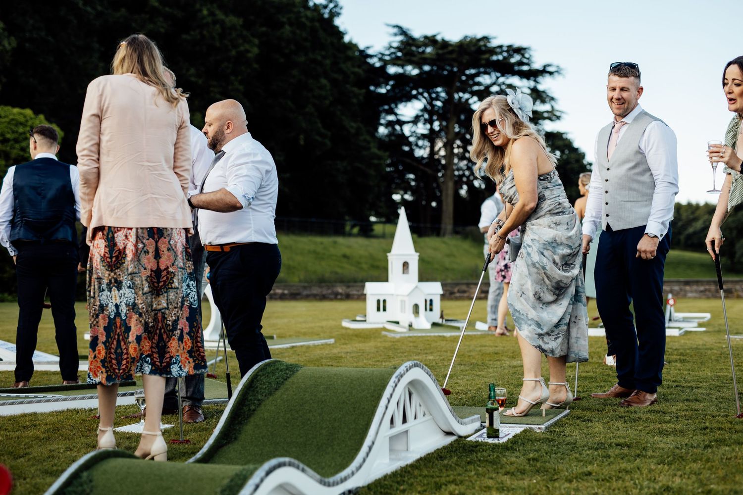 Wedding guests playing miniature golf on a lawn with a decorative white church structure.