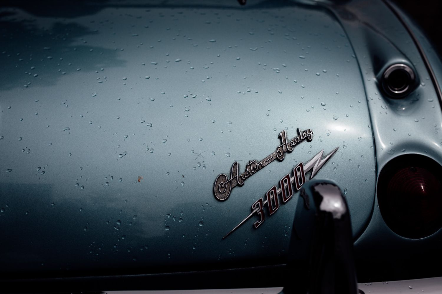 Raindrops on sleek silver car surface with classic script lettering visible.
