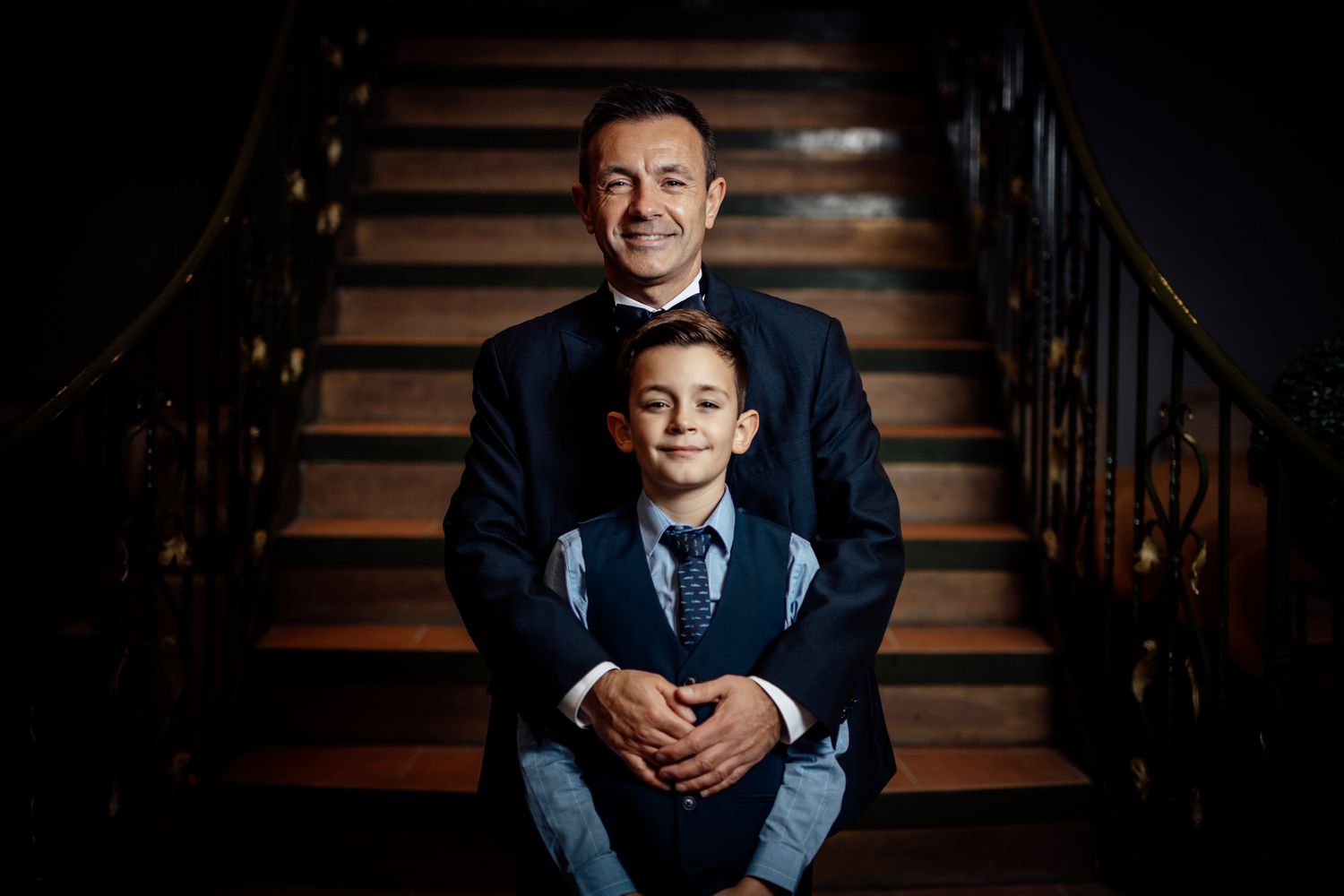 A formal portrait taken on wooden stairs with elegant lighting and dark background.