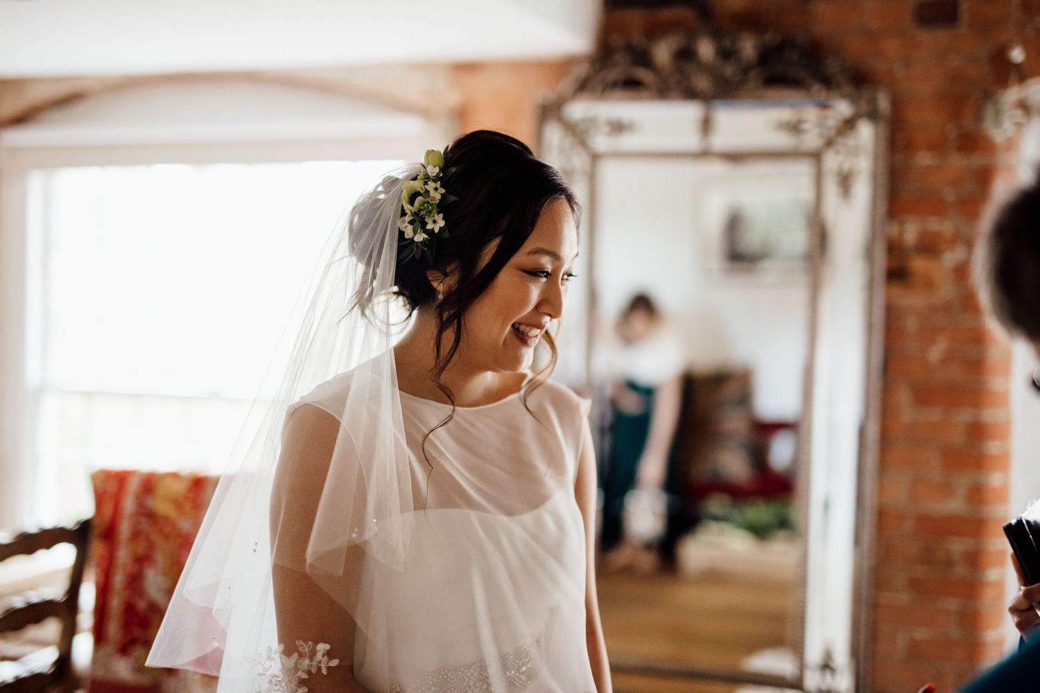 Bride in white veil smiling joyfully in sunlit room with exposed brick walls.