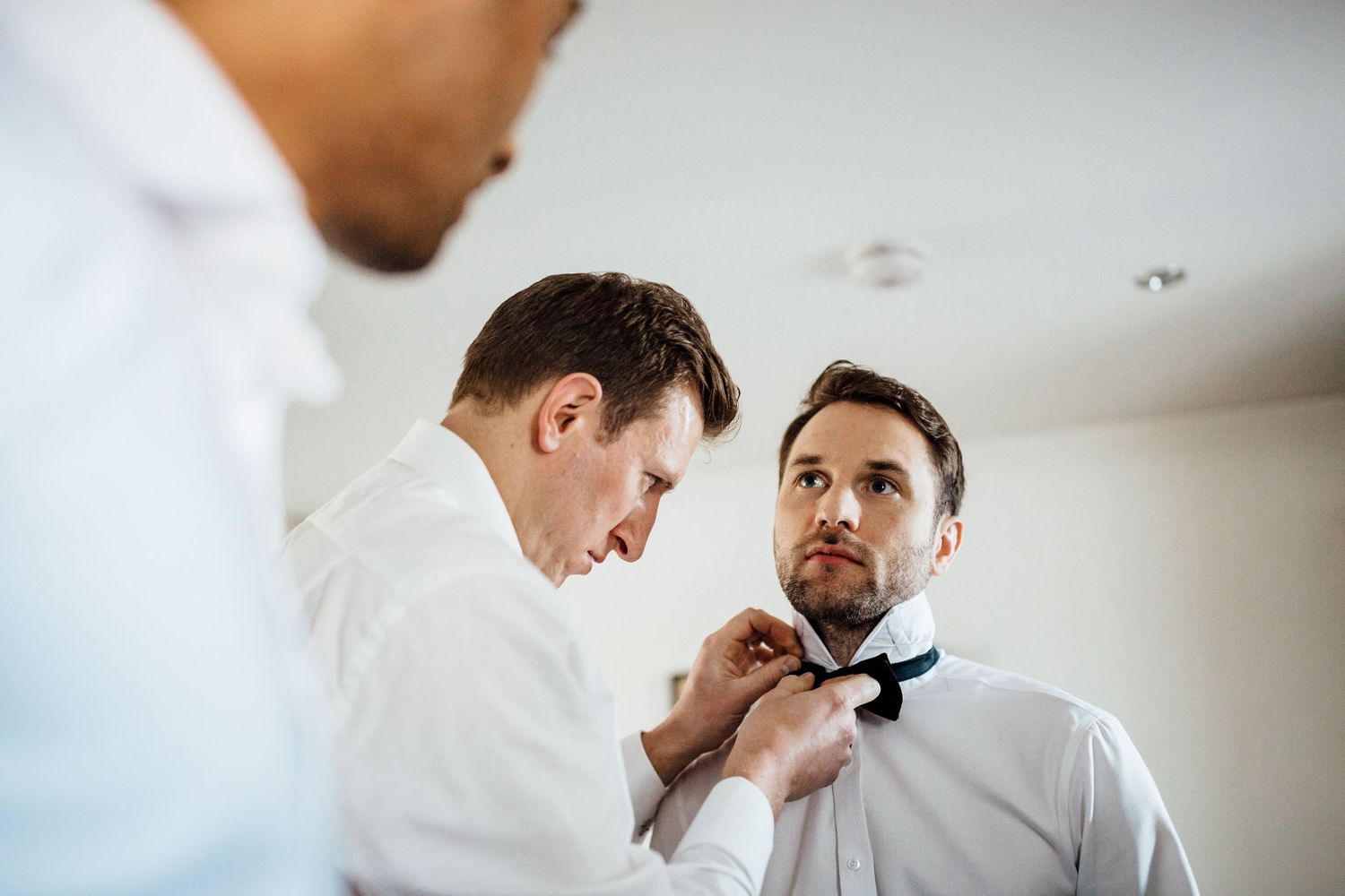 Two people in white dress shirts help adjust a black bowtie during formal preparation.