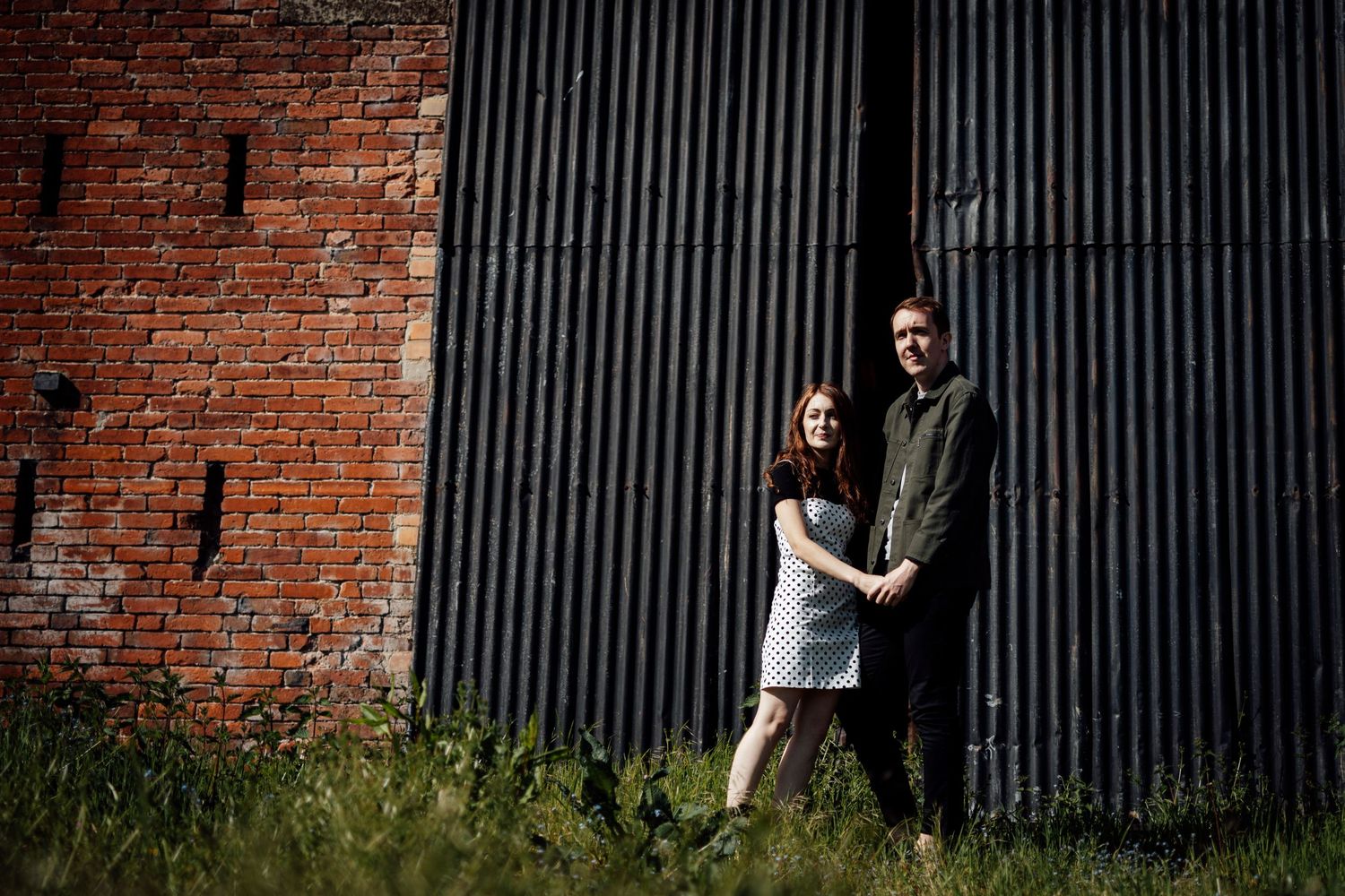 A couple poses against a rustic brick wall with vertical metal bars.