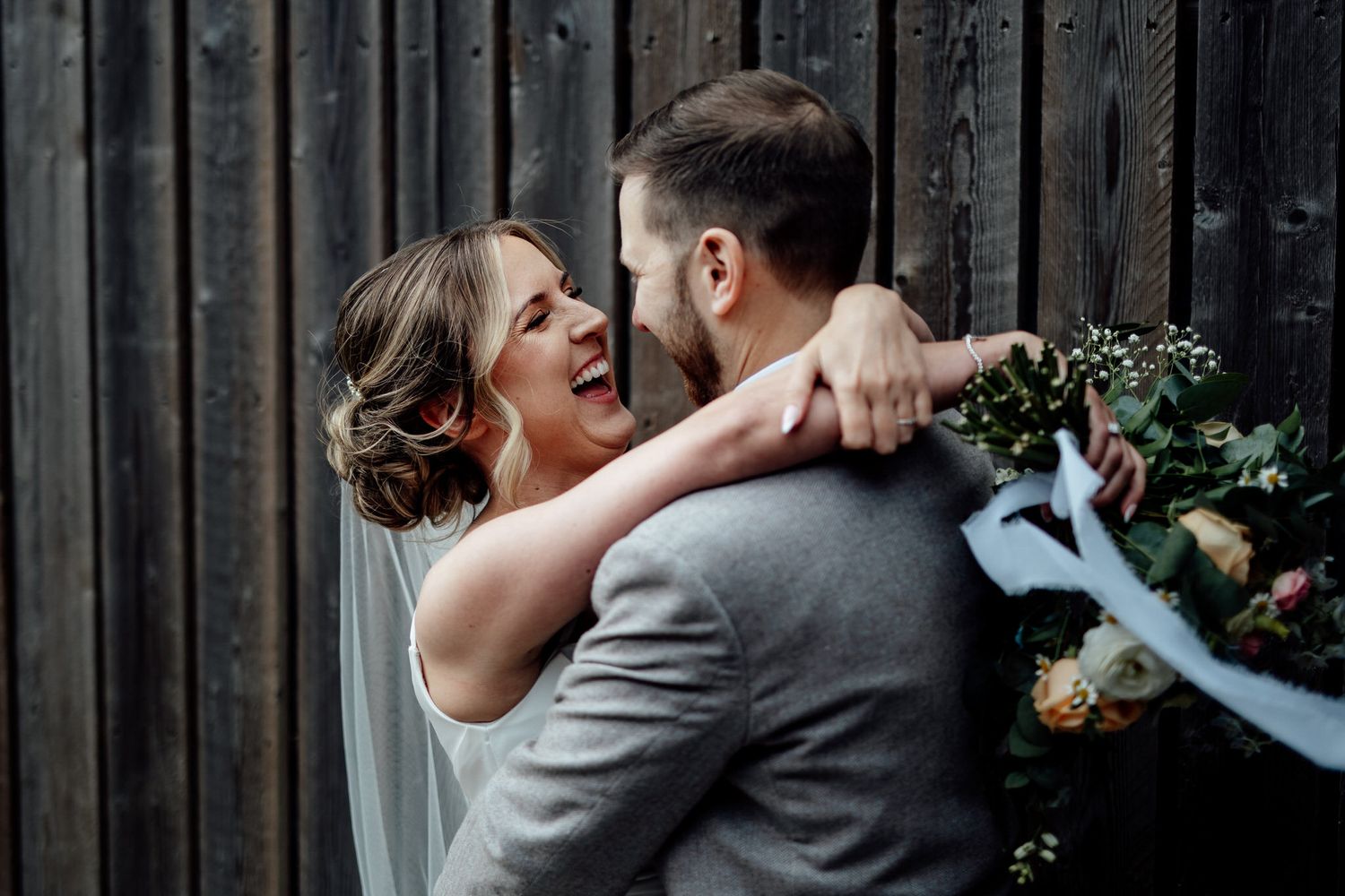 A joyful wedding moment captured against a rustic wooden wall backdrop with greenery and floral decorations.