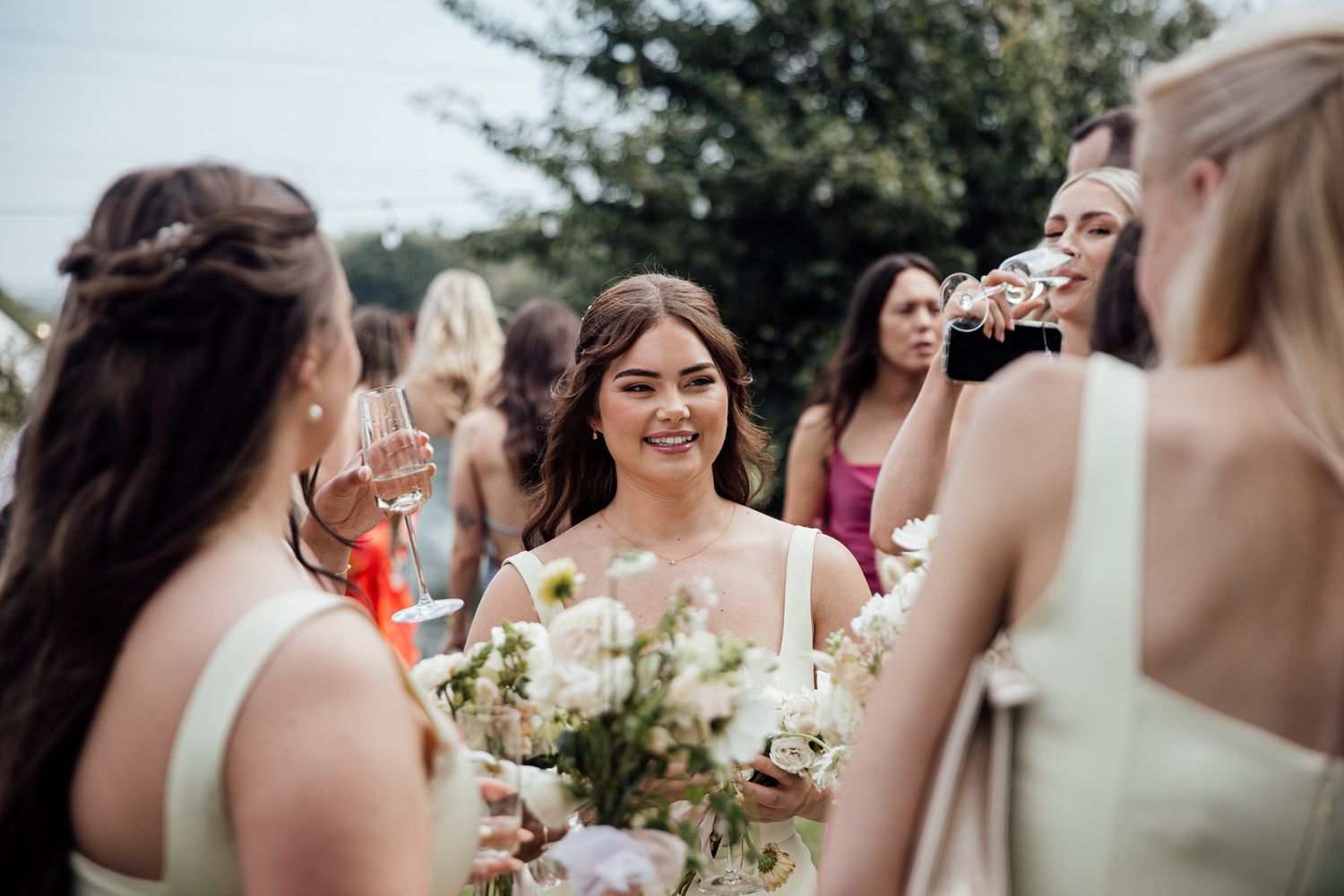 Wedding party members in light dresses gather outdoors for candid moments.