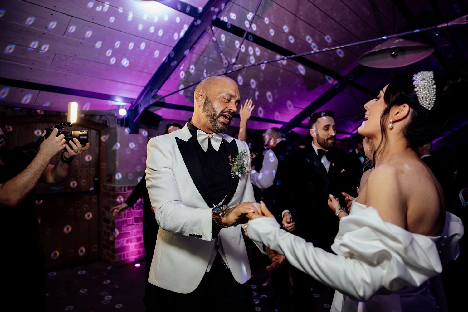 Wedding guests dancing and celebrating under vibrant purple lighting at reception venue.