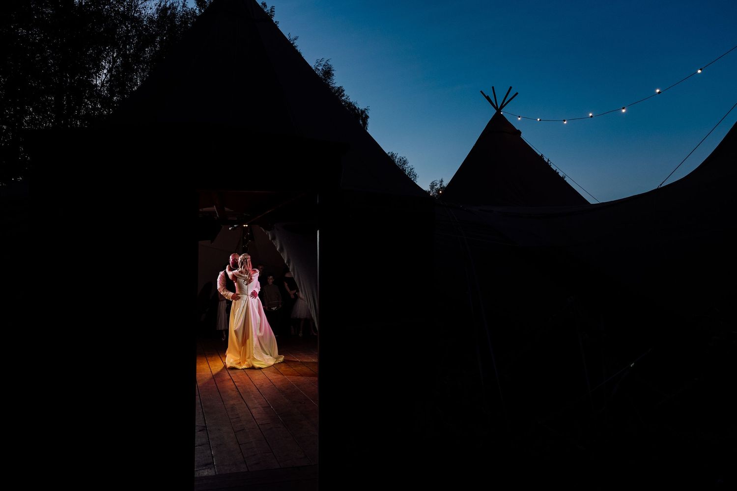 Dramatic nighttime wedding portrait with couple illuminated in doorway beneath string lights and teepee.