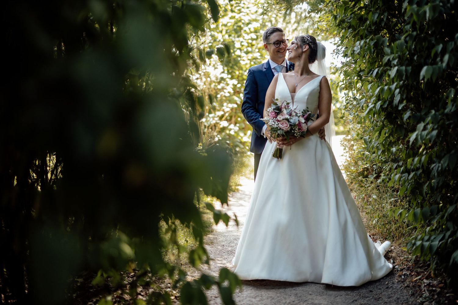 Wedding couple embraces in a garden setting with natural sunlight filtering through trees.
