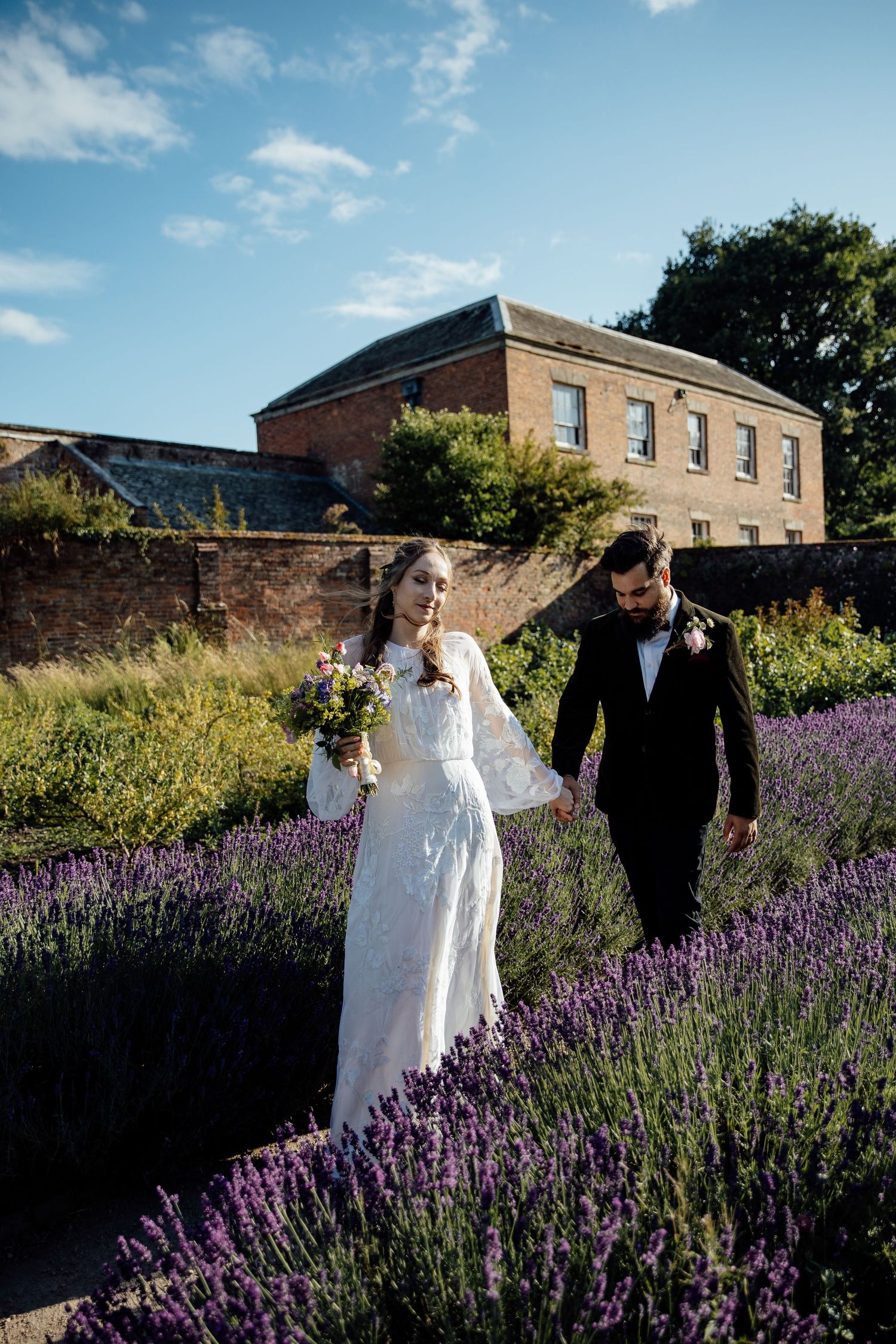 A couple strolls through a beautiful garden with blooming purple lavender and brick buildings in the background.