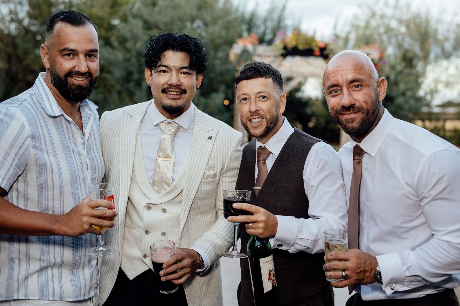 Four well-dressed men smiling and holding drinks together outdoors at a wedding celebration.