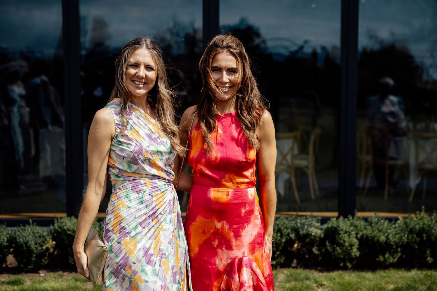 Two guests in colorful summer dresses pose together on a sunny outdoor lawn.