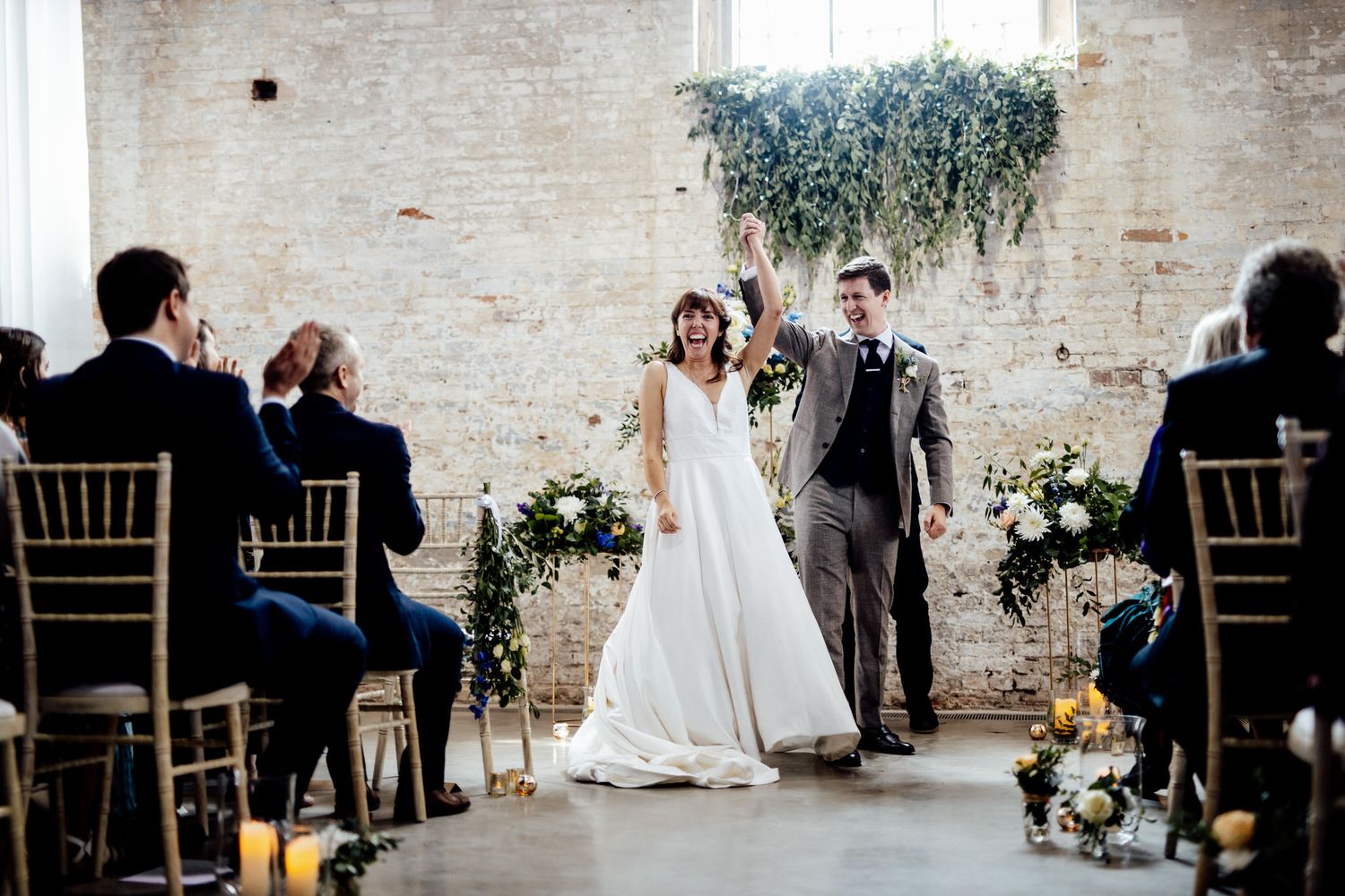 Wedding guests seated in rows of gold chiavari chairs inside an industrial brick venue space.