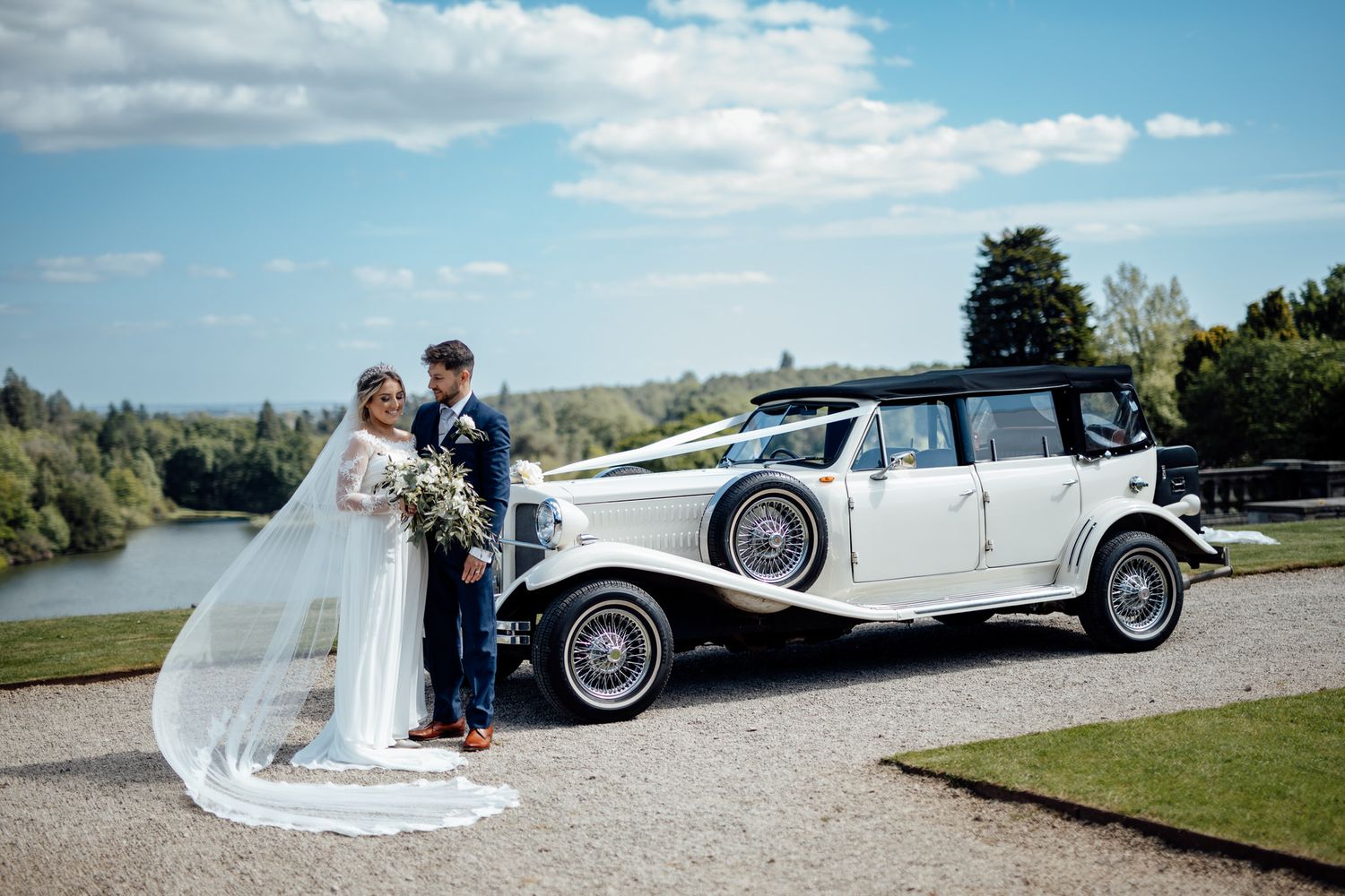 Newlyweds posing beside classic white vintage car on scenic driveway.