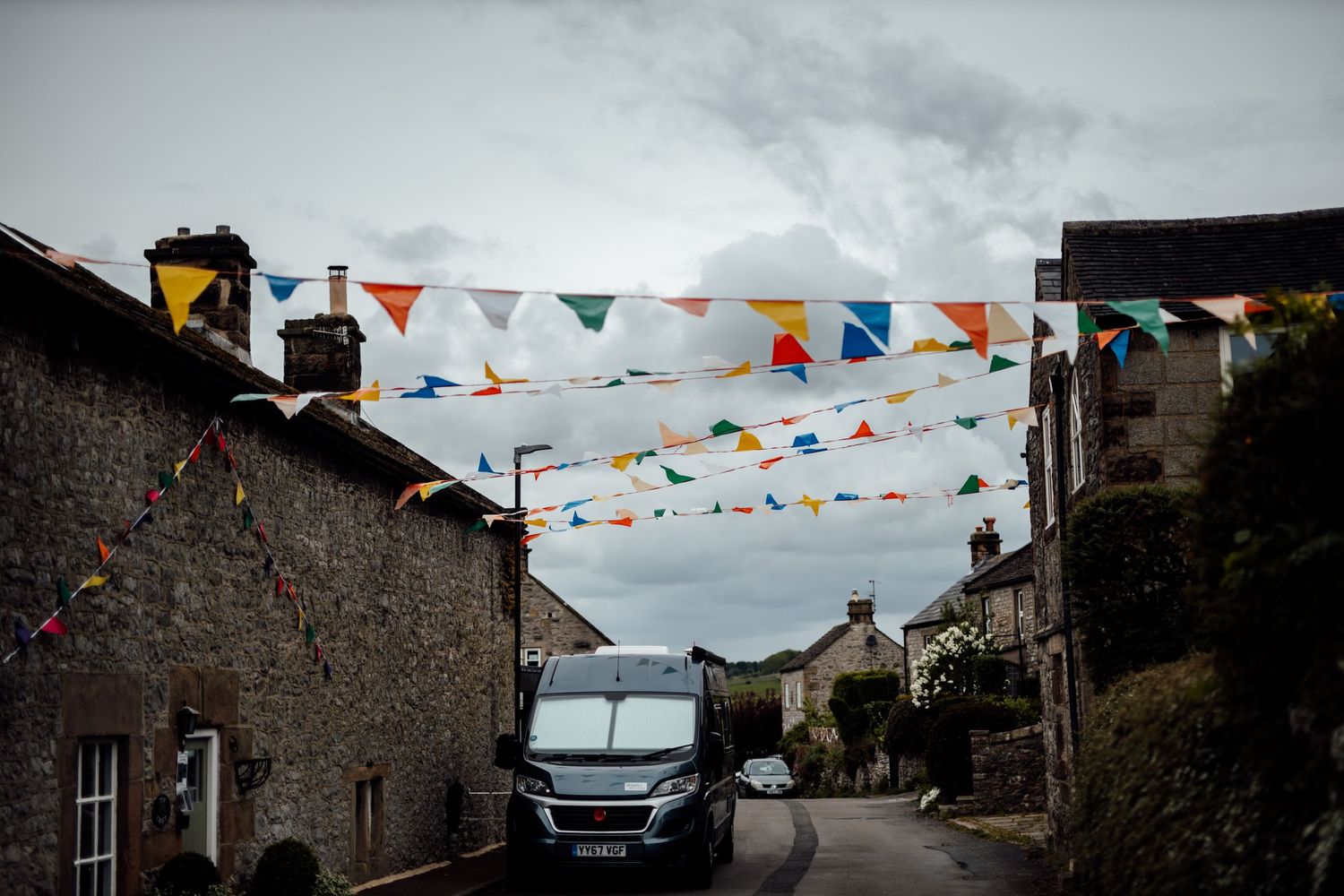 A village street decorated with colorful bunting flags against a cloudy sky.