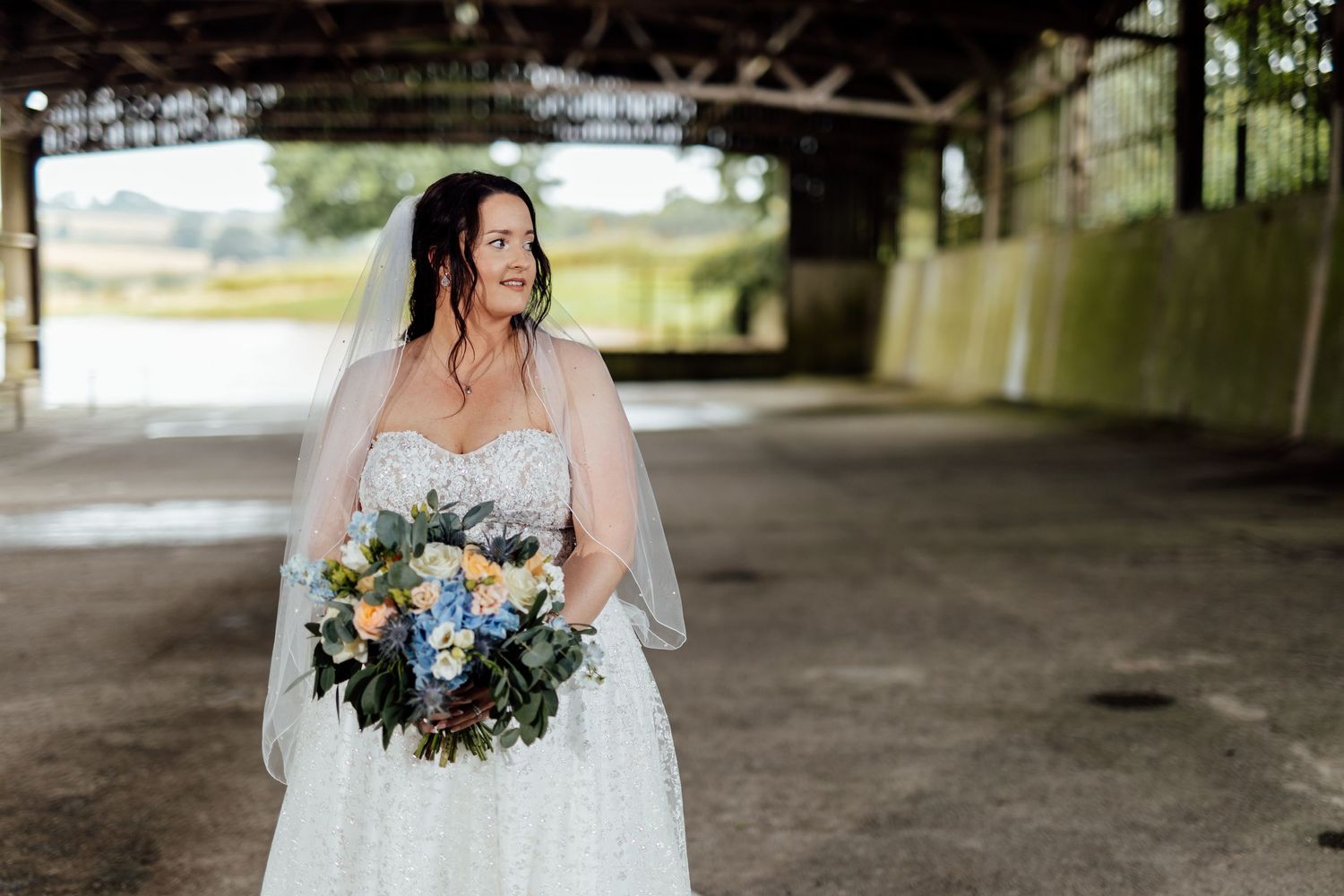 Bride in white lace dress holds vibrant blue and white bouquet under concrete bridge.