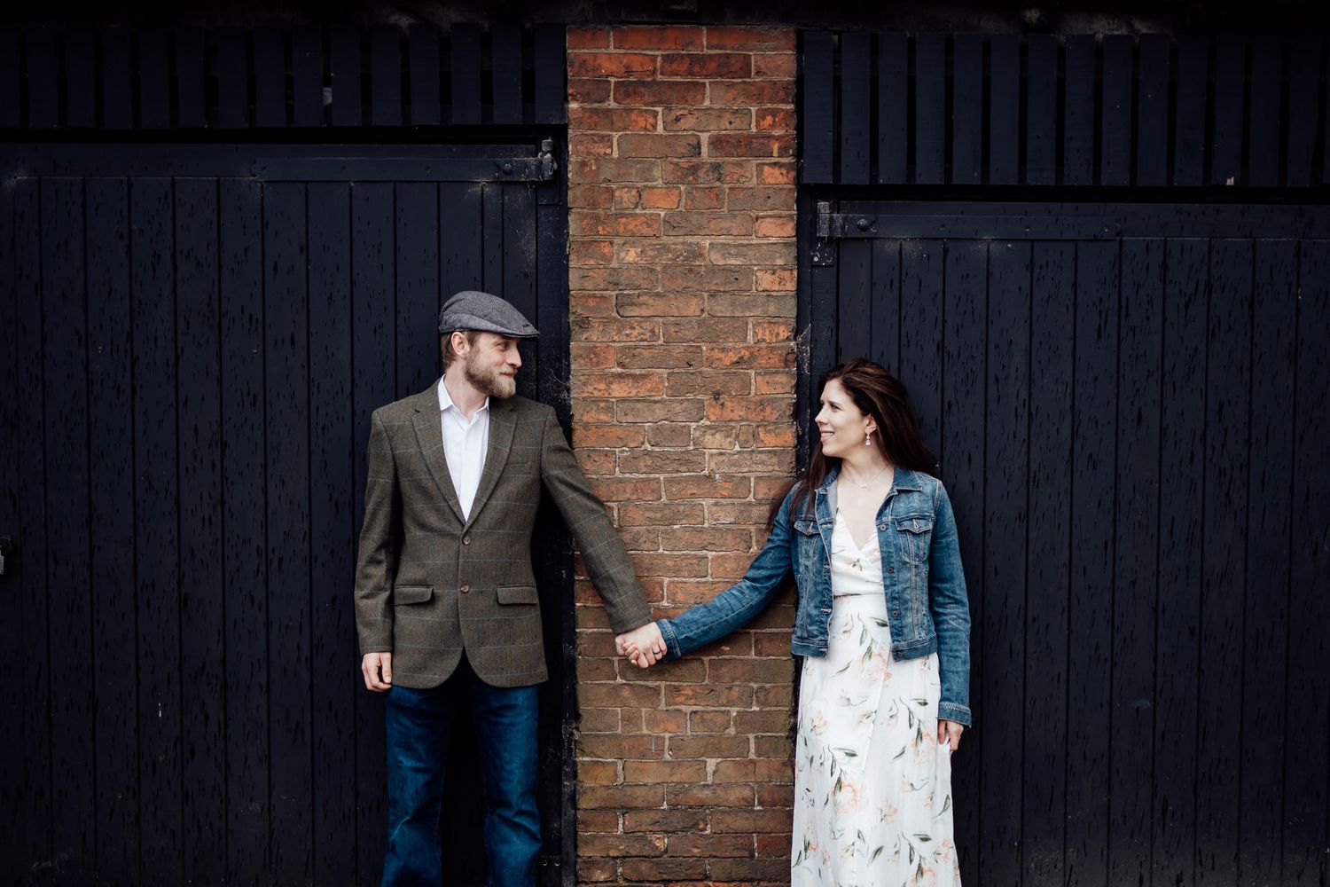 A couple stands against a dark wooden door and brick wall backdrop in casual attire.