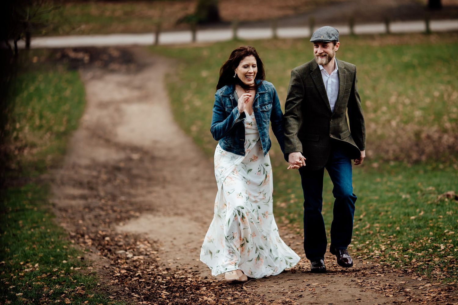 A couple walks along a dirt path in autumn, with her wearing a lace dress and denim jacket and him in a blazer and cap.