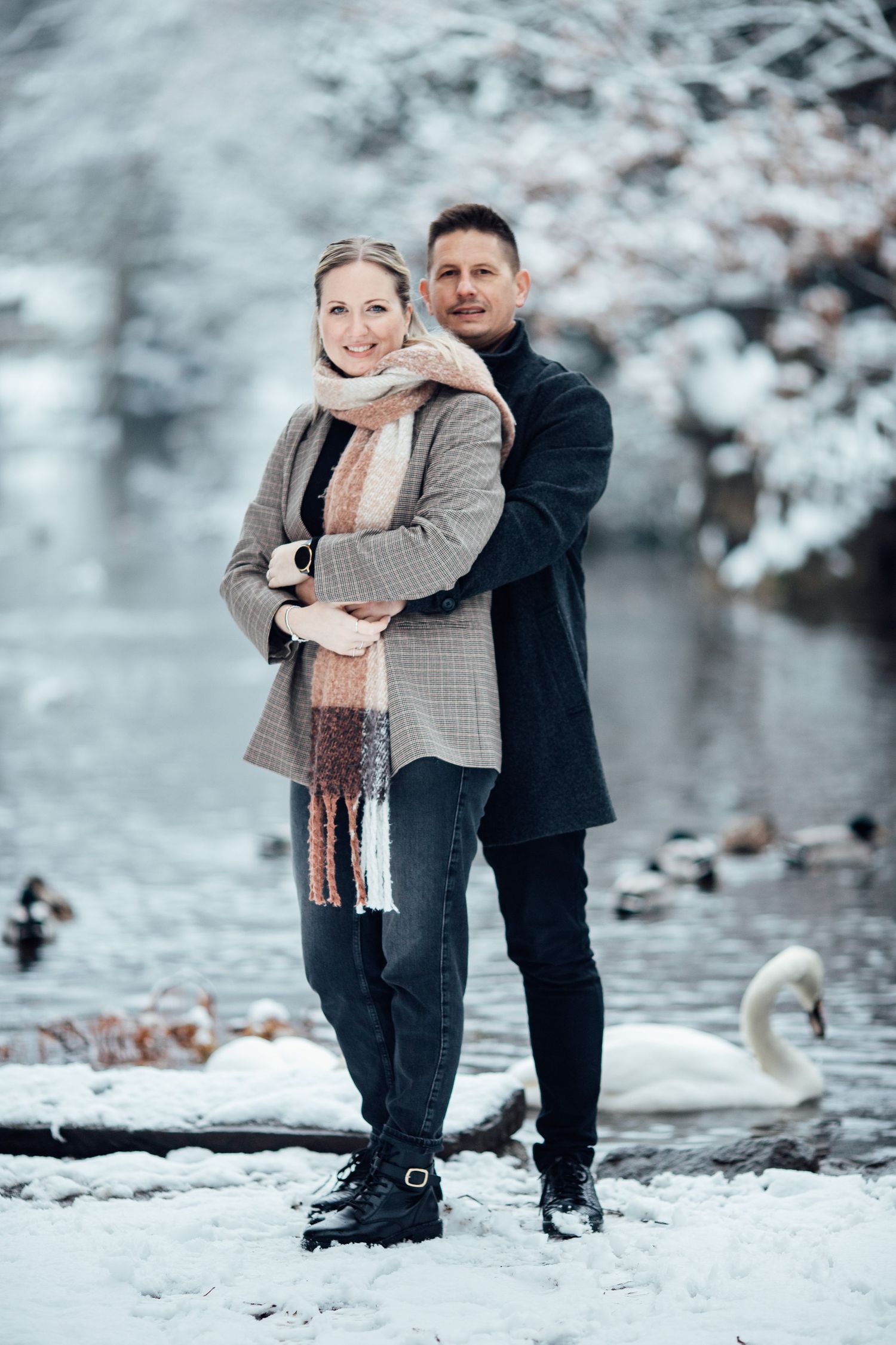 A tender moment captured by a pond in winter with snow-covered trees in the background.