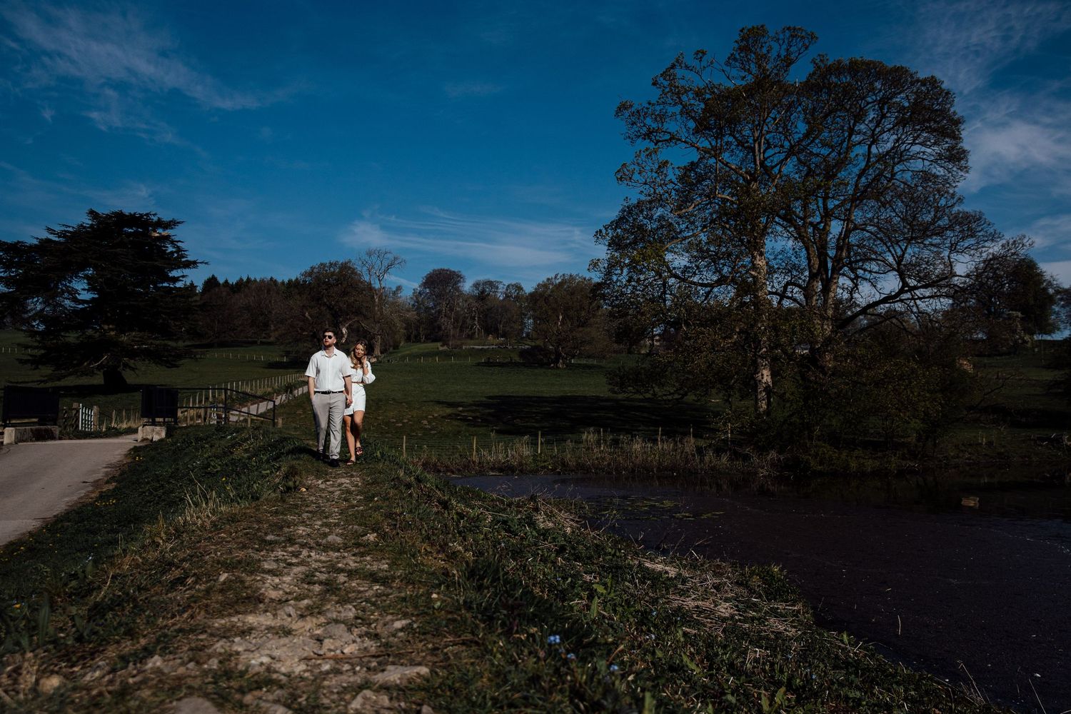 A couple walks along a winding path with blue skies and trees in the background.
