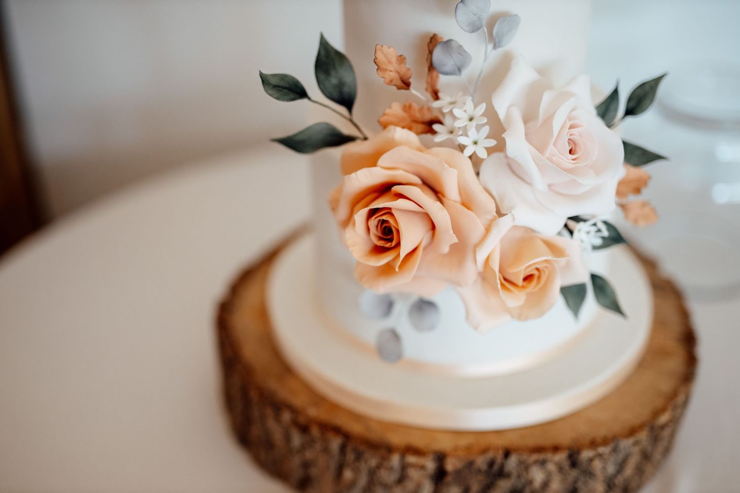 Wedding cake decorated with delicate peach roses and white flowers on wooden slice.