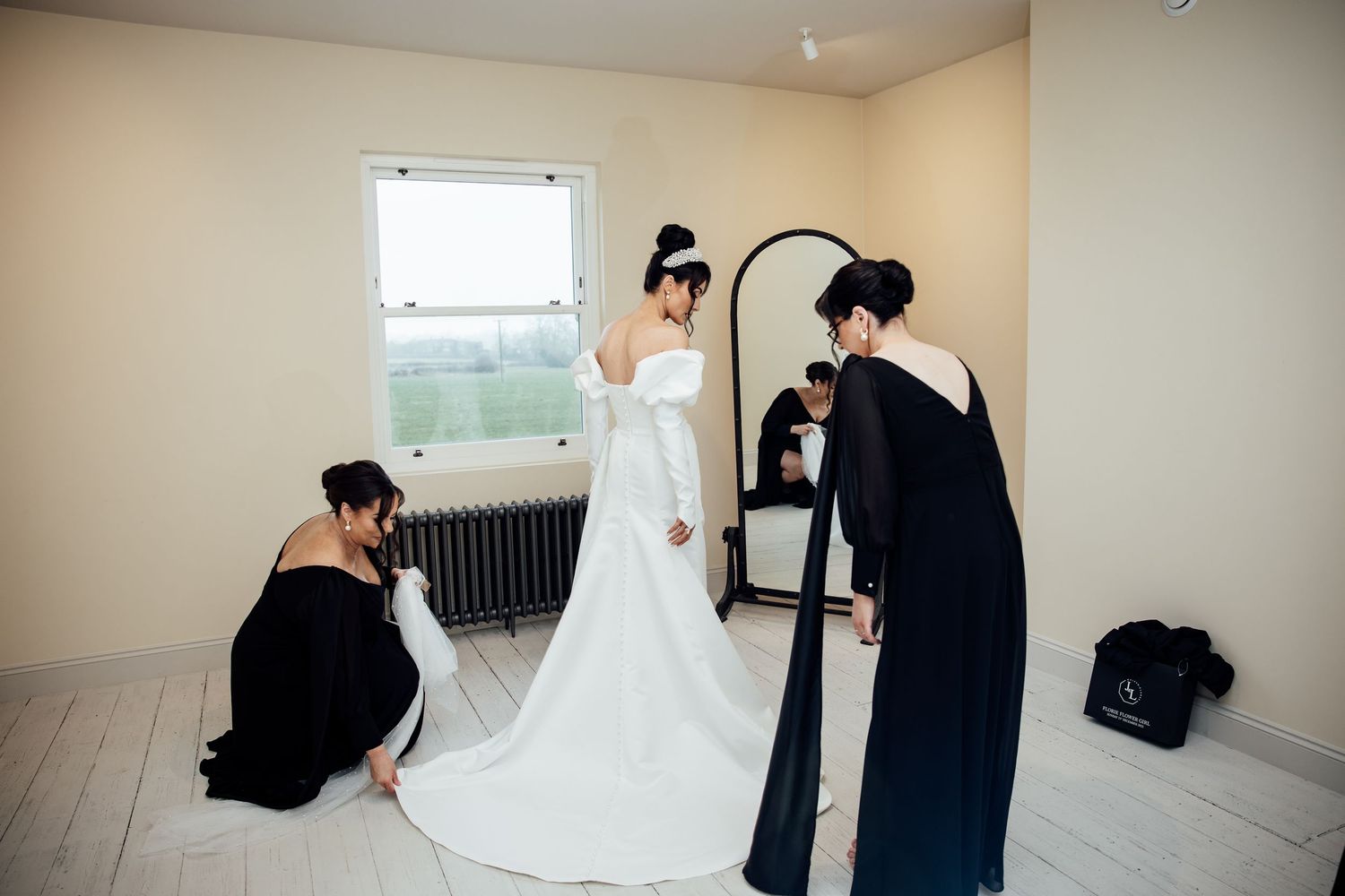 Wedding dress preparation scene with attendants helping adjust the gown in a bright room.