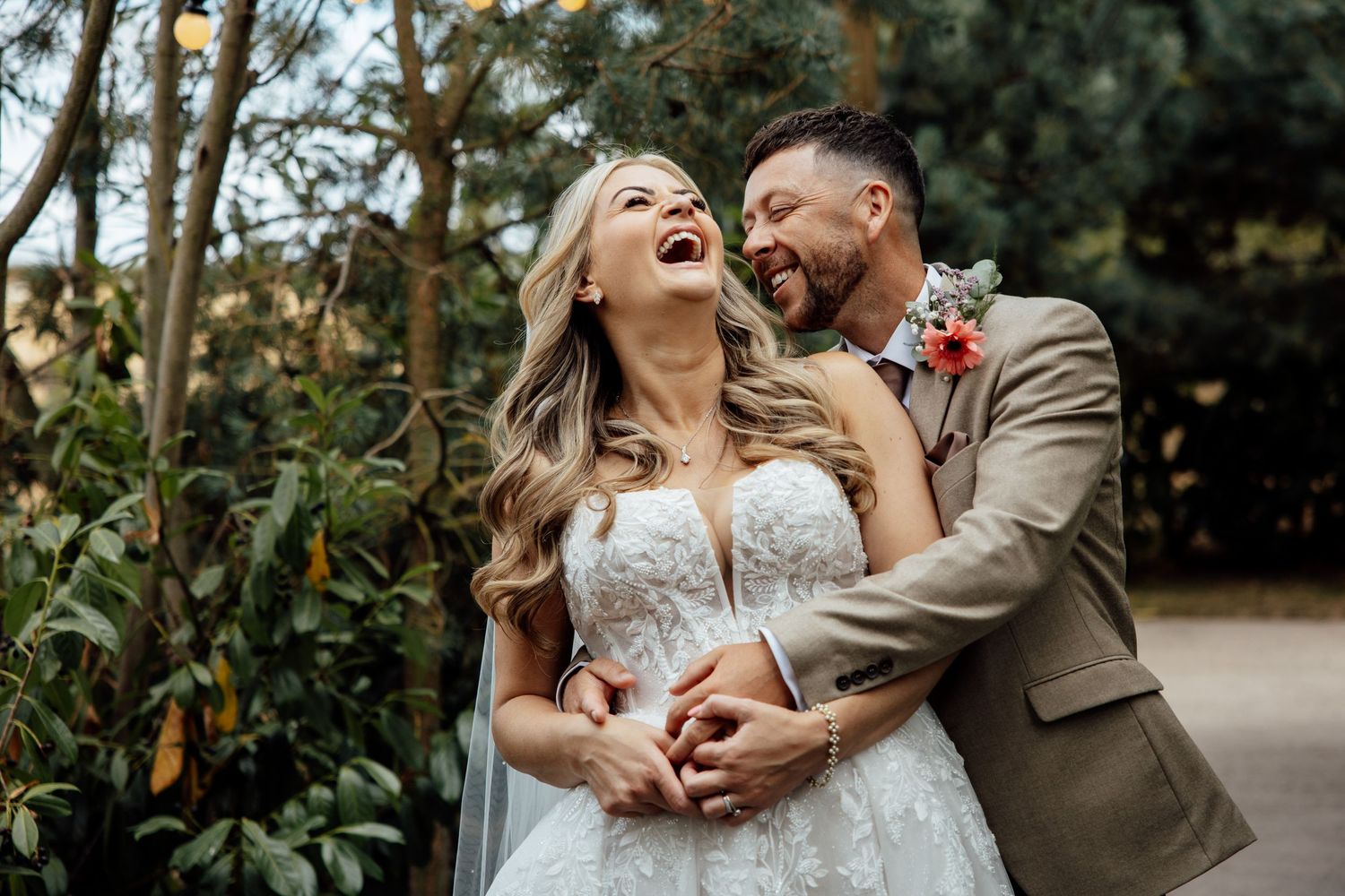 Joyful bride in lace gown laughs as groom in tan suit embraces her in garden setting.