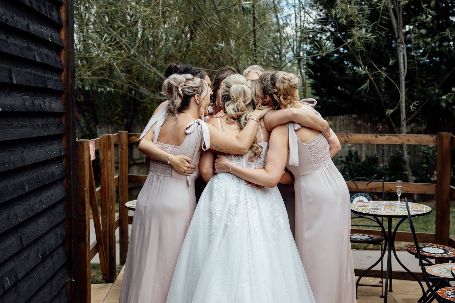 Bride in white gown sharing a heartfelt group hug with bridesmaids in blush pink dresses outdoors.