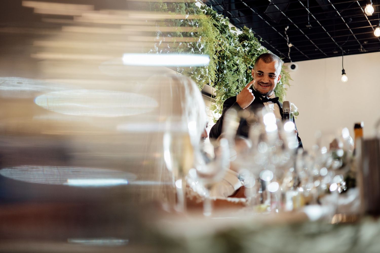 Smiling wedding MC in bow tie gesturing at a beautifully set reception table with glassware.