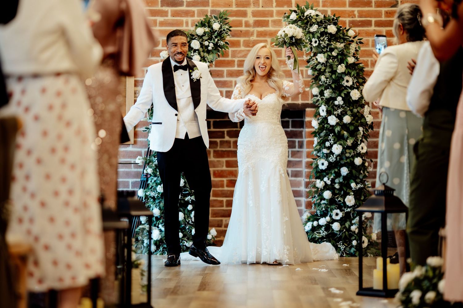 Newlywed couple celebrating at the altar in a brick-walled venue adorned with white floral arch decorations.