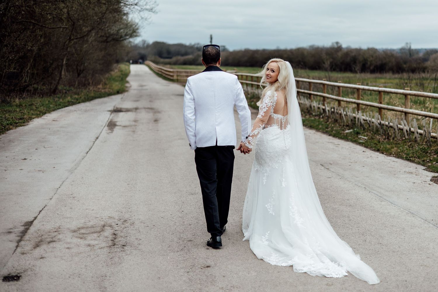 Bride and groom walk hand in hand down a scenic road, bride wearing elegant lace gown with veil.