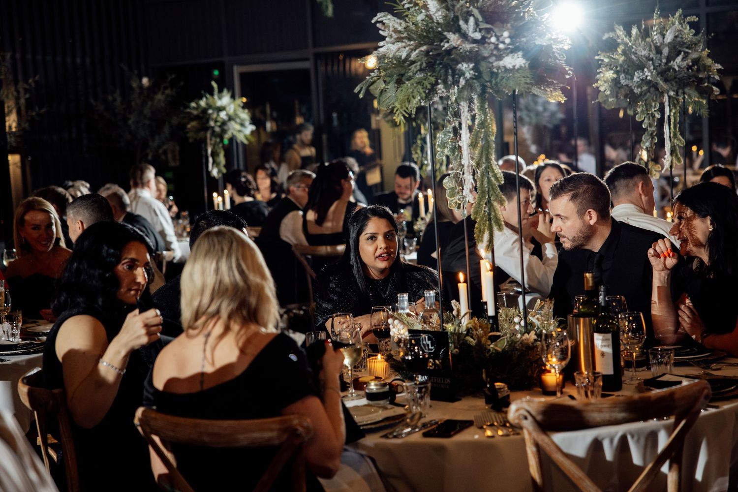 Wedding reception dinner setup with elegant table settings and greenery in a dimly lit venue.