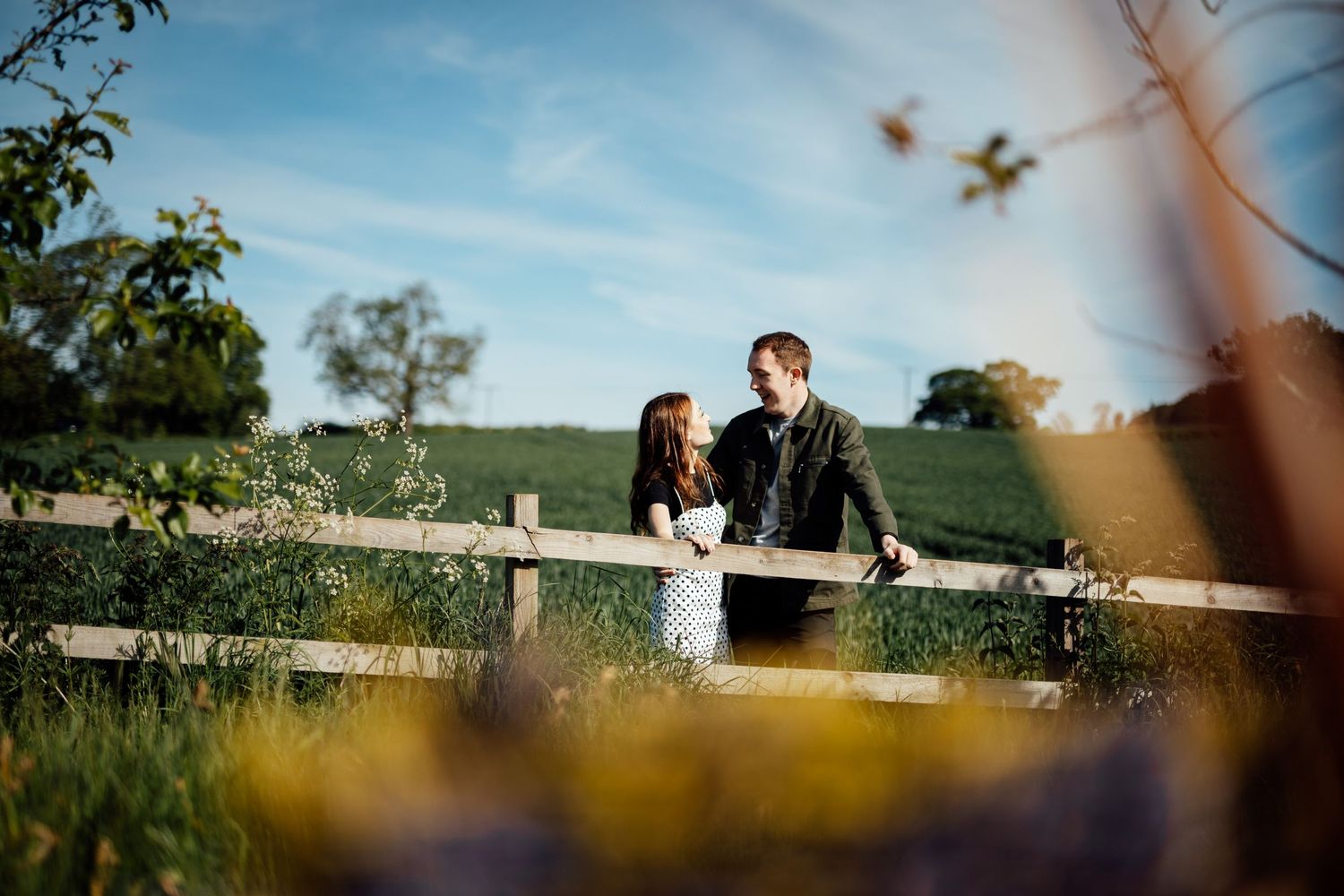 Couple sharing a romantic moment by a rustic wooden fence in natural sunlight.